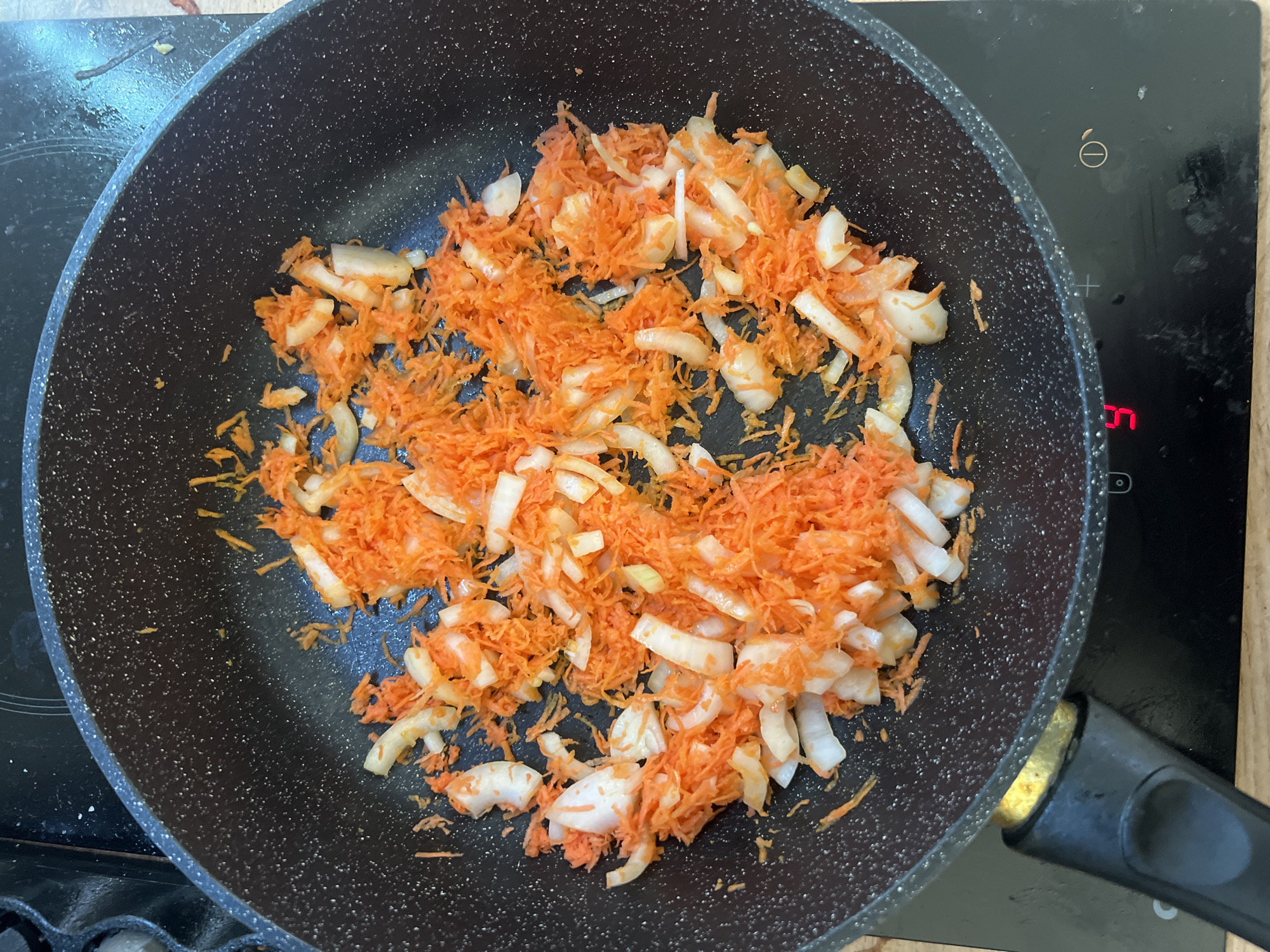 The image shows a close-up, overhead view of a black frying pan containing shredded carrots and chopped white onions. The pan is on a black electric stovetop, which has a digital display showing the number "5" in red. The stovetop also shows symbols for power and heat settings. The frying pan has a black speckled interior and a black handle. The scene is indoors, and based on the available information, this image does not provide any specific location details within the city of Saint Petersburg, Russia, or any other city. No people are visible in the image. The food appears to be raw and in the process of being prepared for cooking.