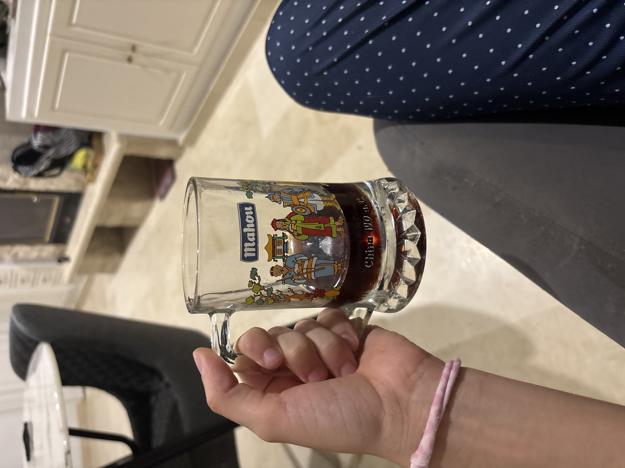 The image shows a close-up of a person's hand holding a glass mug filled with a dark liquid, likely beer. The mug has a decorative design featuring figures in historical attire and the word "Mahou," a Spanish beer brand. The scene appears to be indoors, with a blurry background that includes furniture, possibly a dining table and chairs, and a cabinet. The floor is tiled. The lighting suggests an indoor setting, possibly a home or restaurant. There is no visible indication of the city of Orihuela, Spain, in the image.