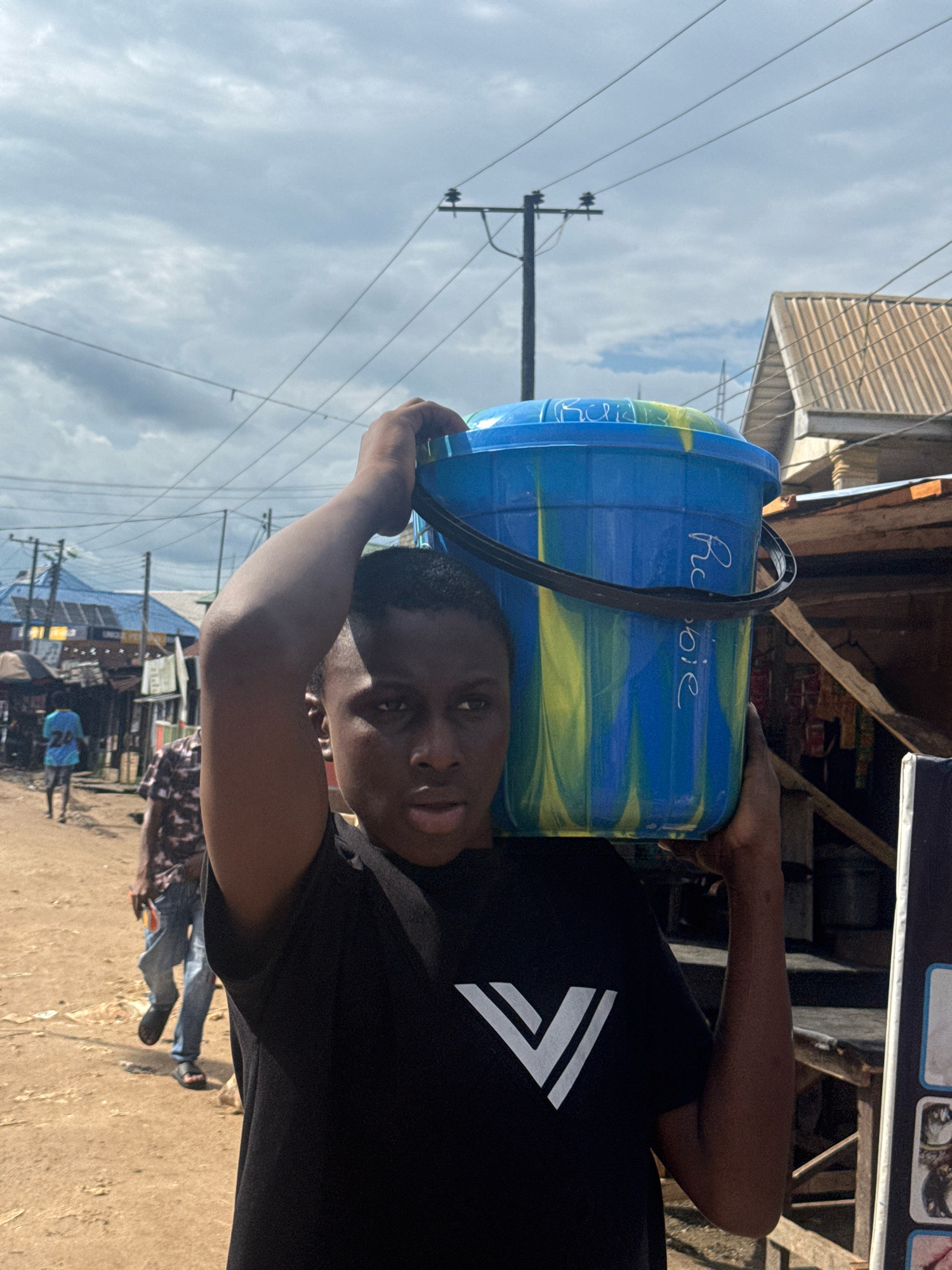 In Ihiagwa, Nigeria, a young individual carries a large blue and yellow swirled bucket filled with water on their head. The person is wearing a black t-shirt with a white V-shaped design. The background depicts a dusty street scene with market stalls, corrugated metal roofs, and utility poles with wires. Other individuals are visible walking in the background, and the sky is cloudy. The scene appears to be a typical day in a bustling Nigerian community.
