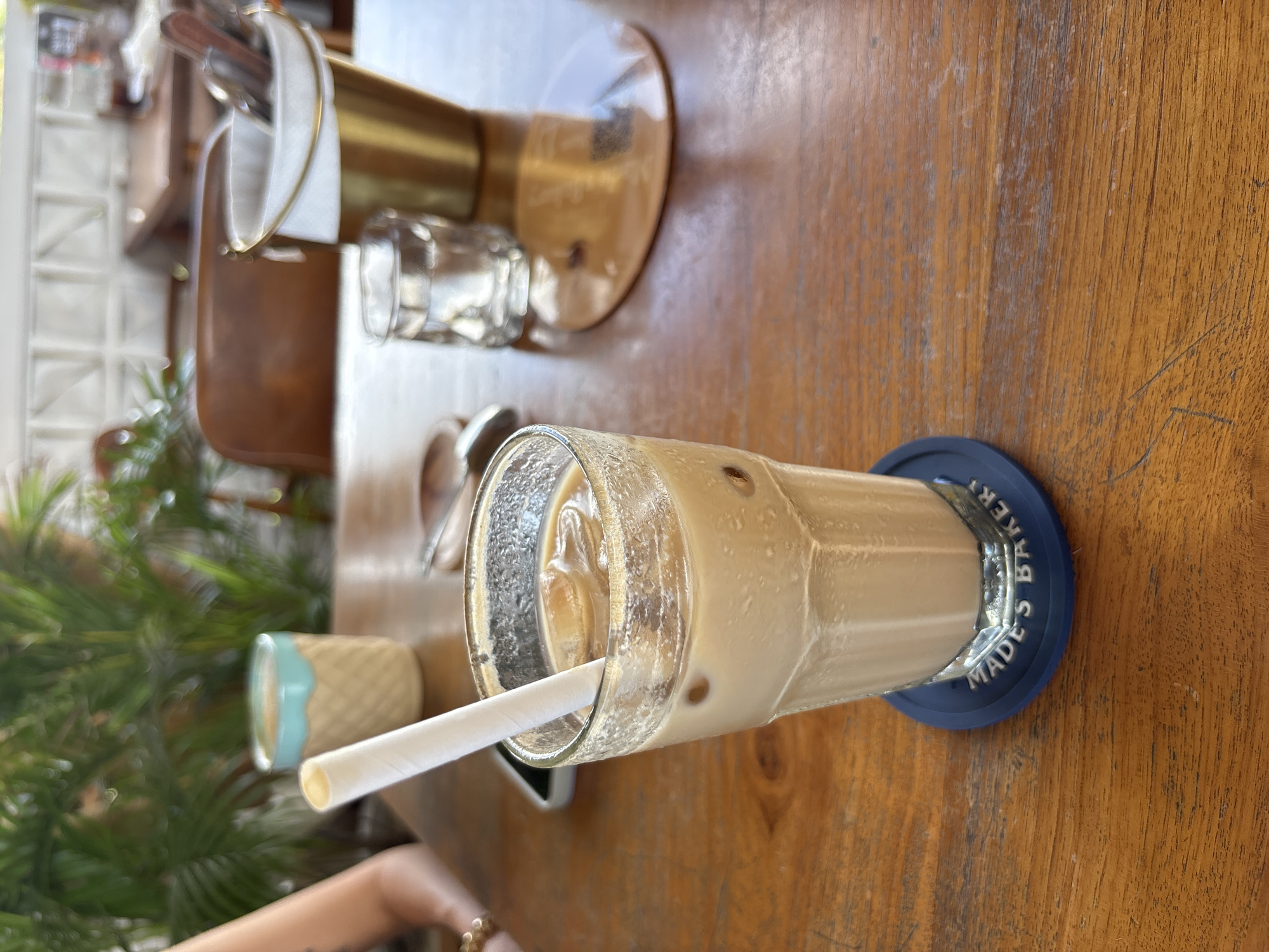 The image shows a glass of iced coffee on a blue coaster that says "Made's Baker" on a wooden table. There's a drinking straw in the glass. In the background, there is a cutlery holder, a small glass, sunglasses, and a small decorative ice cream cone. There's greenery visible in the lower-left corner and part of a person's arm and jewelry. The location is likely Made's Baker in Kuta Selatan, Indonesia.