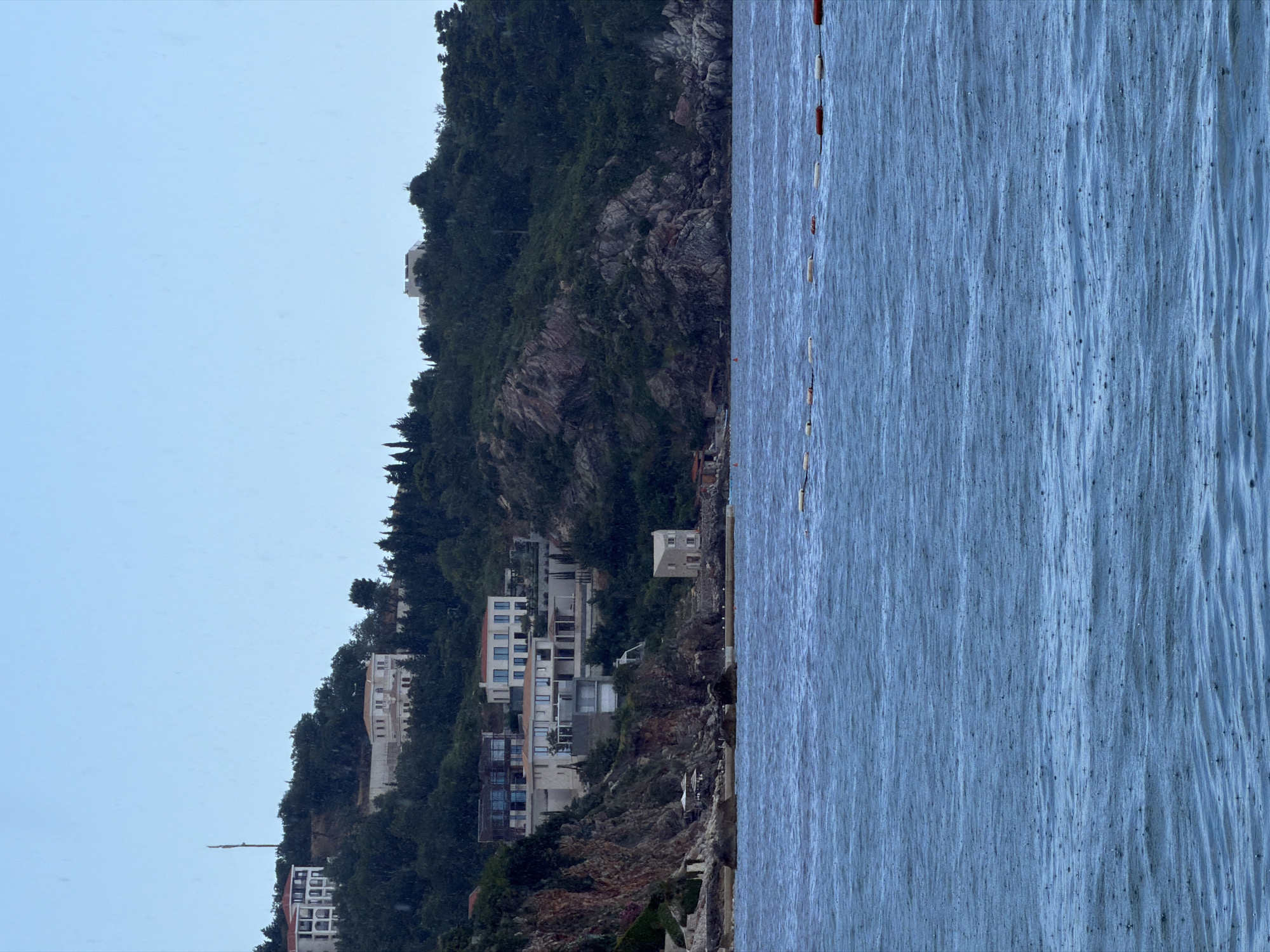 This media file depicts a seaside view of buildings nestled on a steep, tree-covered mountainside overlooking a body of water. The scene is oriented vertically, with the water occupying the right side of the frame and the landmass on the left.
The buildings appear to be modern, with multiple stories, balconies, and large windows. They are situated at various levels on the rocky incline, surrounded by lush green vegetation. Some structures are more contemporary, with clean lines and white or light-colored facades, while others show signs of age or appear to be under construction or renovation. At the very bottom of the mountain, some smaller structures and a pier or retaining wall are visible near the water's edge.
The water itself is a deep blue with visible ripples and texture, suggesting it is a calm sea or ocean. A line of buoys, some red and some white, stretches across the water near the shore, possibly marking a swimming area or a boundary.
The sky above is a pale, overcast blue, indicating a diffused light, likely early morning or late afternoon, or a cloudy day. There are no people visible in the frame, and the overall mood is serene and quiet. The presence of the buildings on the mountainside suggests a coastal settlement or resort area in Drobnići, Montenegro. There is no visible text in the image.