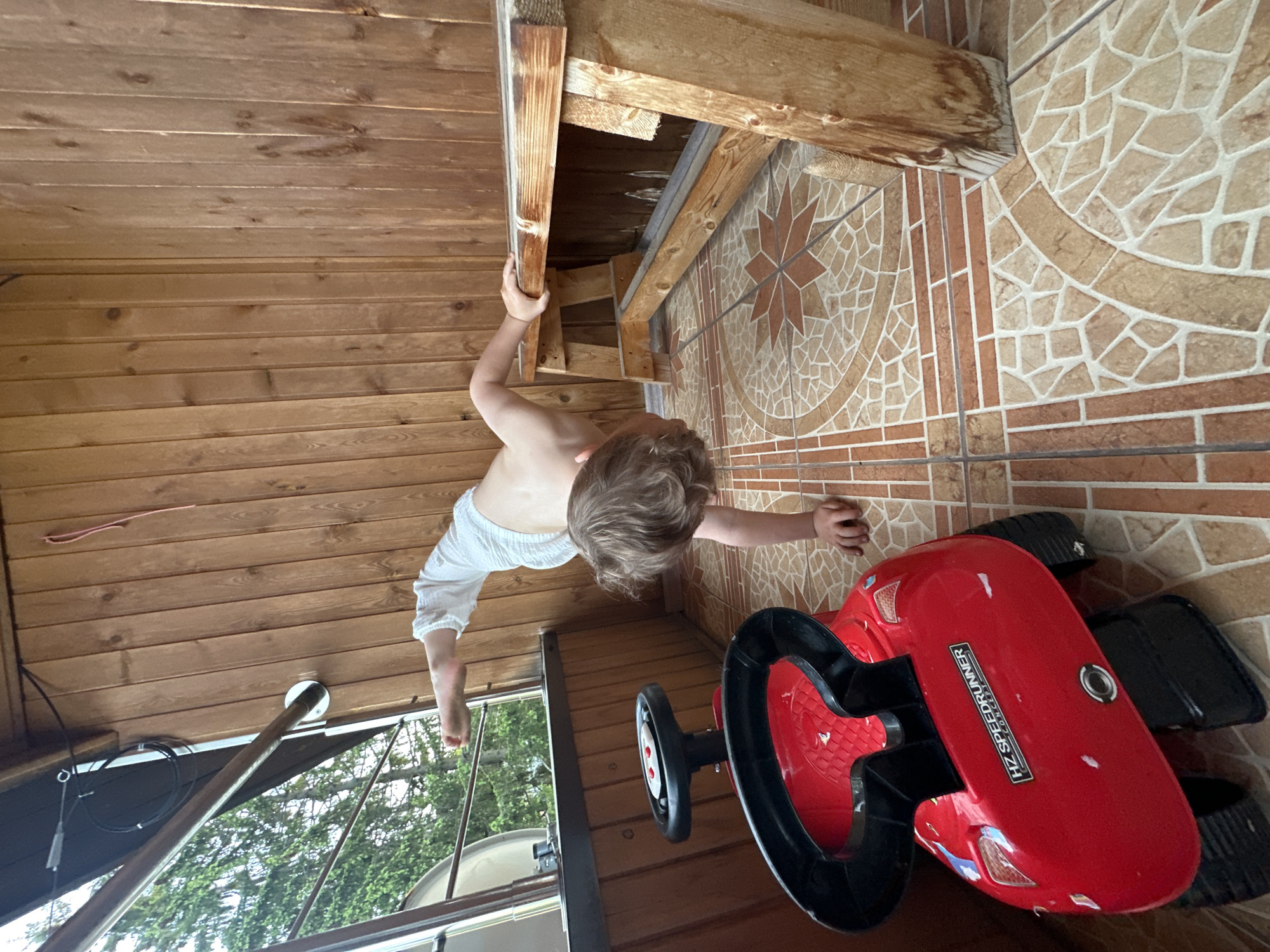 A child wearing white clothing is hanging from a wooden structure in what appears to be a covered outdoor area, possibly in Nizhny Novgorod, Russia. The child's hand is gripping a wooden beam. A red toy car is on the tiled floor. The background shows wood paneling.