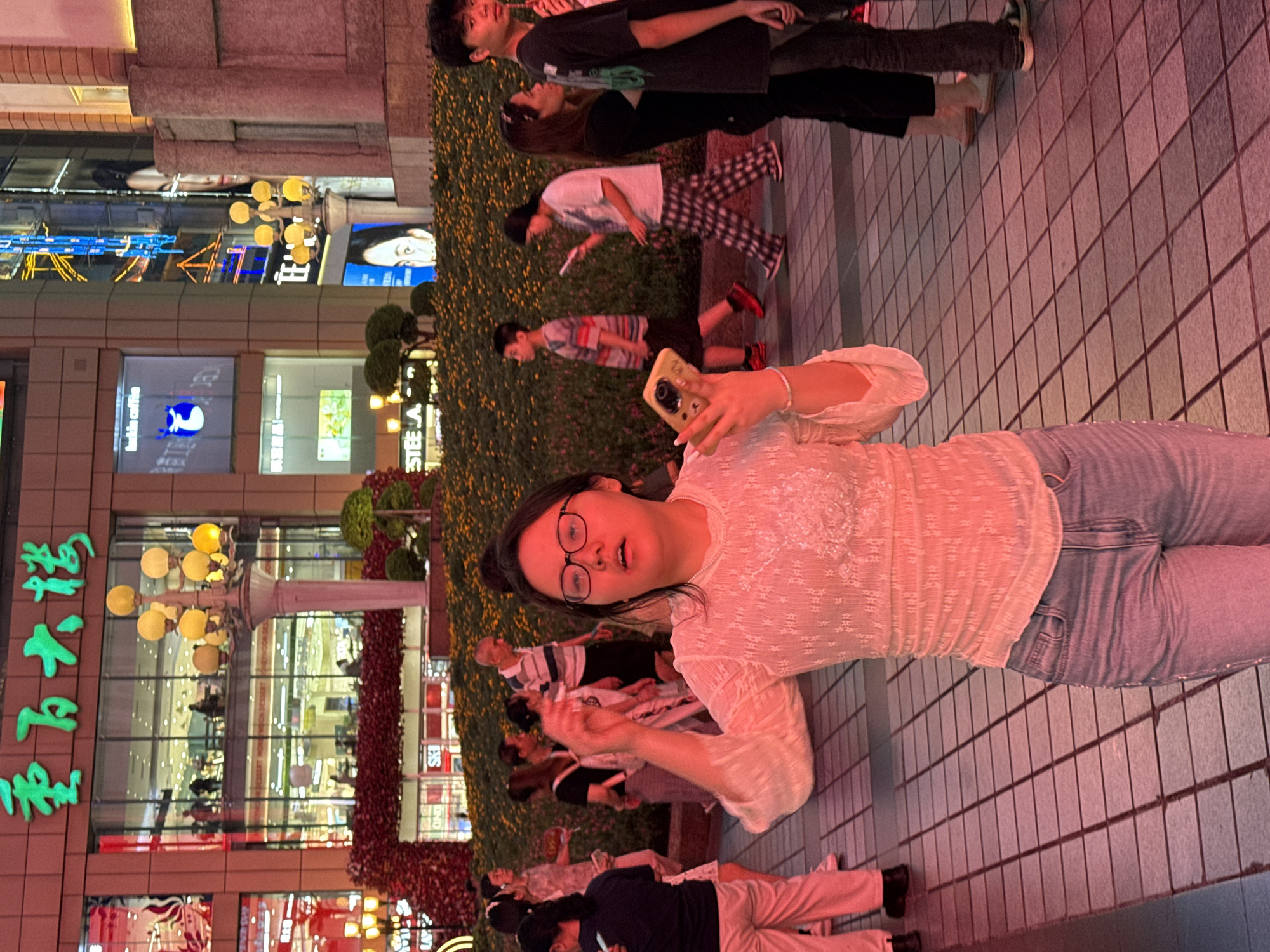 In the city of Yuzhong District, China, a young woman with dark hair and glasses stands in the foreground, looking towards the camera. She is wearing a sheer, long-sleeved white blouse and light-wash jeans. She holds a yellow smartphone in her right hand, her arm extended. In the background, several other people are walking or standing, with a large building featuring illuminated signage and red floral decorations. The ground is paved with square tiles, and the scene is lit with warm, artificial lighting, creating a reddish-pink hue.