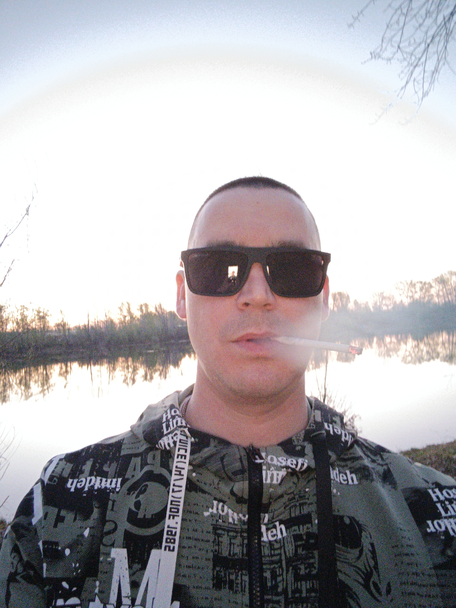 A medium-shot selfie of a Caucasian man with a shaved head and dark sunglasses, facing the camera. He is smoking a cigarette. He is wearing a green, black, and white patterned hoodie with words printed on it and a white lanyard around his neck. Behind him is a body of water reflecting trees on the opposite bank, consistent with the landscape of Chaikovsky, Russia, which is known for its proximity to the Kama River and associated bodies of water. A faint halo effect is visible in the sky.