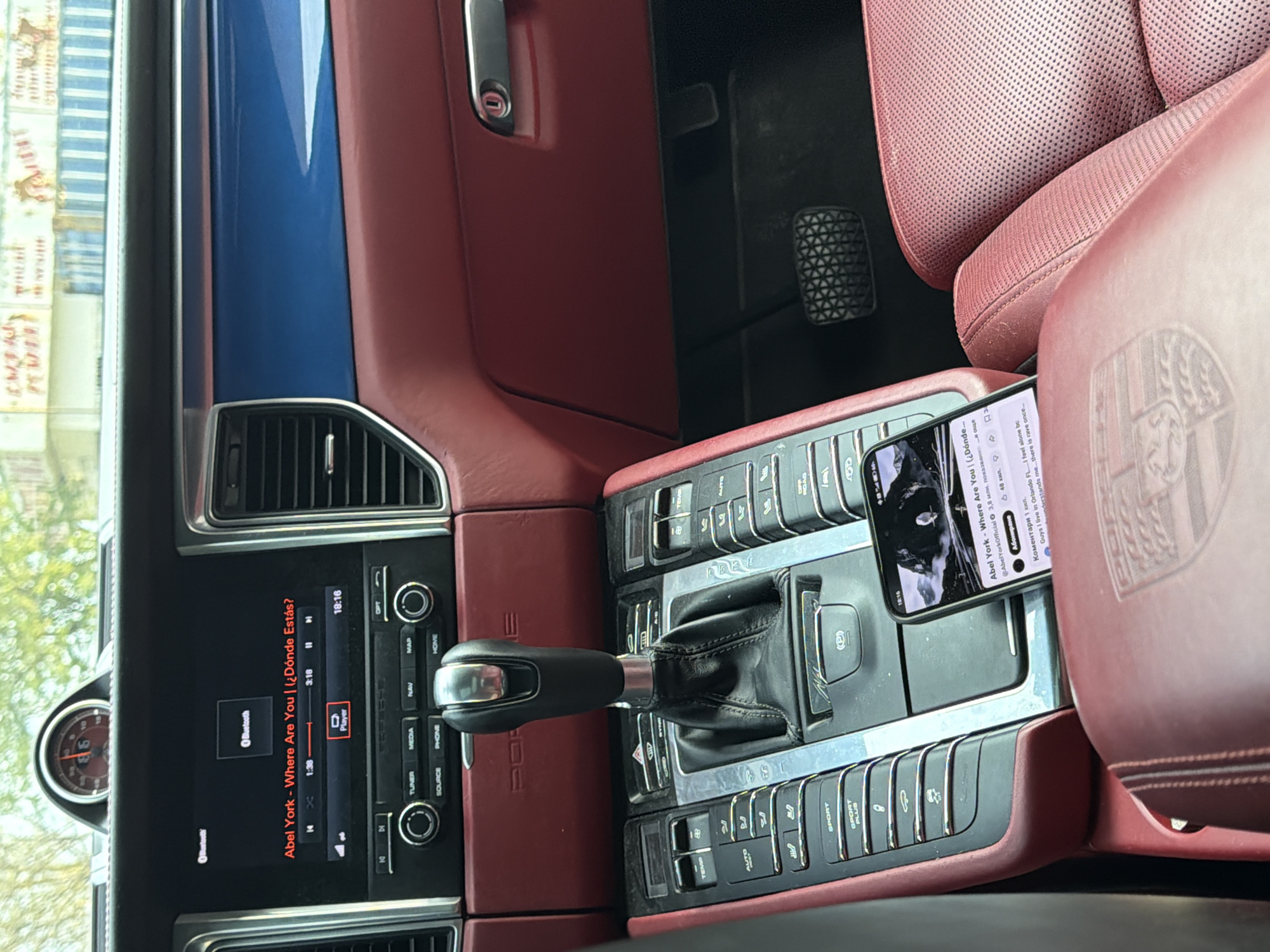 The image captures the interior of a car, likely a Porsche Macan, featuring a luxurious red/burgundy leather interior with a blue trim accent visible on the door panel. The primary focus is on the dashboard and center console, where a smartphone is resting.
The car's infotainment system is active, displaying "Abel York - Where Are You | (¿Dónde Estás?)" playing via Bluetooth. The song is 1 minute and 38 seconds into a 3-minute, 18-second track. The car's clock and the phone both show the time as 18:16, indicating early evening. The dashboard also features an analog clock, partially visible, which seems to confirm the time around 6 o'clock.
The center console is rich with controls, including buttons for navigation, media, phone, tuner, and various car settings like "SPORT" and "SPORT PLUS." The gear shifter is prominently placed in the center.
The smartphone screen displays a YouTube interface, showing the same song "Abel York - Where Are You | (¿Dónde...)" with 3.8 million views and 48 thousand (likely likes or subscribers). Visible comments include one in English: "Guys I live in Orlando FL....I feel alone bc understands me...there is rave once...". Other text on the phone screen is in Bulgarian, including "показвания" (views), "Абониране" (Subscribe), and "Коментари" (Comments).
A close-up of the red leather seat shows an embossed Porsche logo. The footwell reveals pedals. The view outside the window is blurred but suggests an urban or suburban setting, consistent with the provided context of Varna, Bulgaria, and the Bulgarian text on the phone.