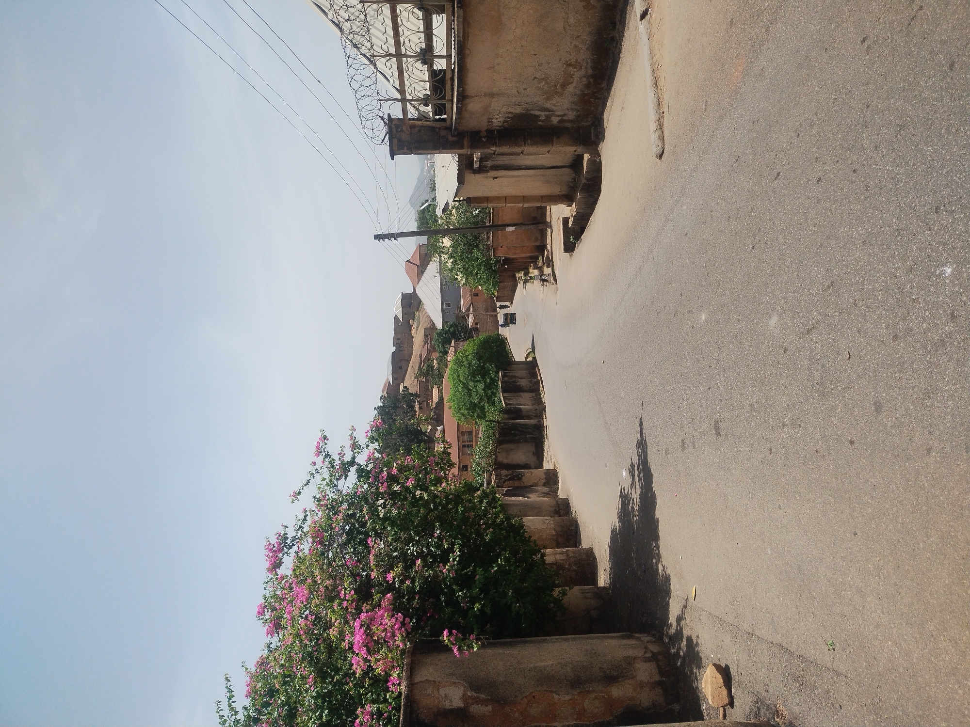 The image depicts a street scene in Jos, Nigeria. The road surface is asphalt and appears weathered. On the left side of the road, a concrete wall supports a large bougainvillea bush with vibrant pink blossoms. Beyond the bush, a set of concrete steps leads up to a building.
On the right side of the road, the concrete wall of a building extends along the street. Above this building, there is a metal security barrier topped with barbed wire. Electrical wires span the sky.
The image is taken at an angle, and the camera is situated higher than ground level, giving a view down the street.