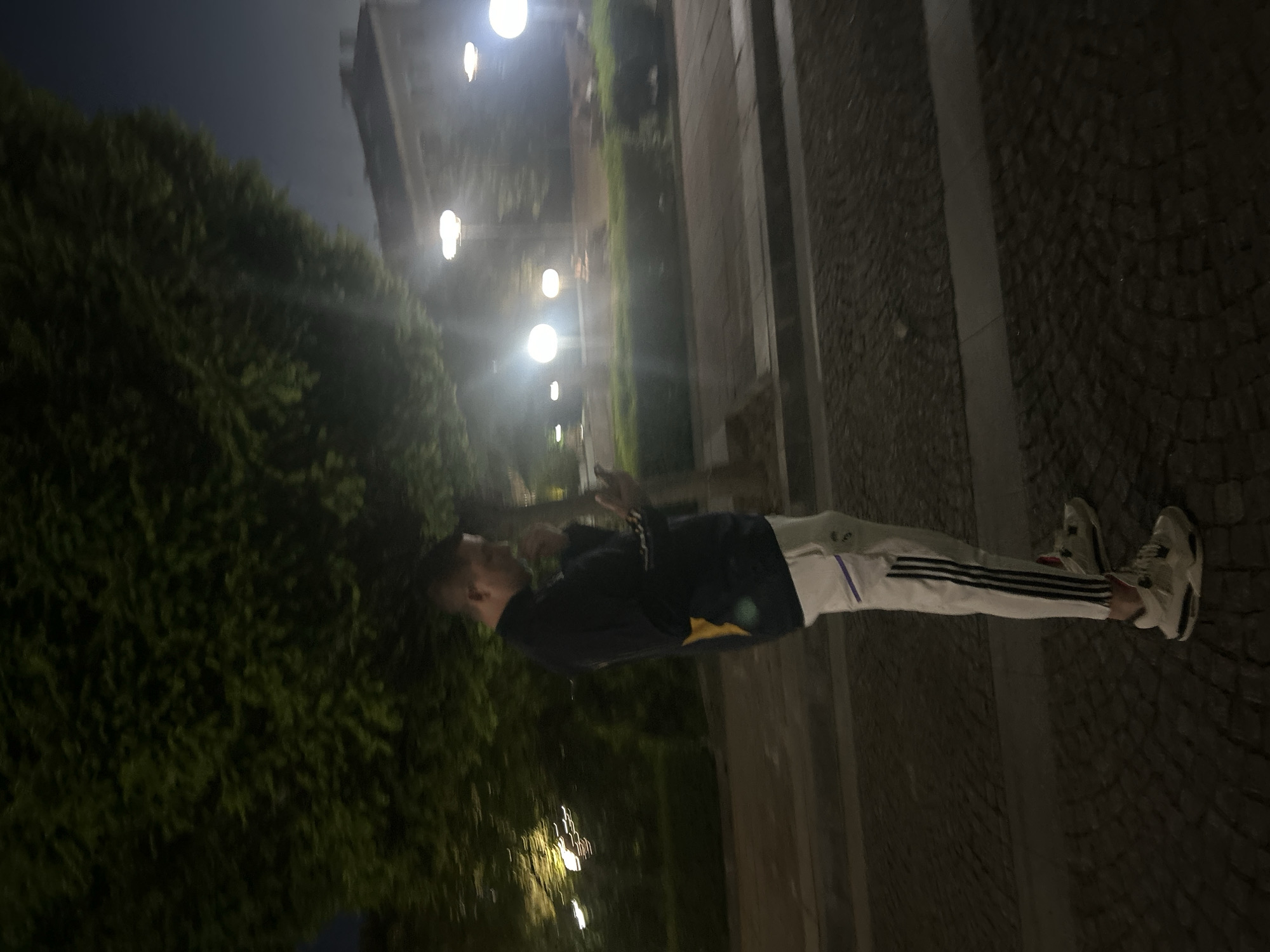A person is standing on a cobblestone walkway next to a building and dense foliage, likely at night. The person is wearing a dark blue sweatshirt with a yellow design, white athletic pants with black stripes, and white sneakers. The person is looking towards the left side of the image and appears to be making hand gestures. Streetlights are visible in the background, suggesting an urban environment. The city of Elhovo, Bulgaria is mentioned as the location of this image.