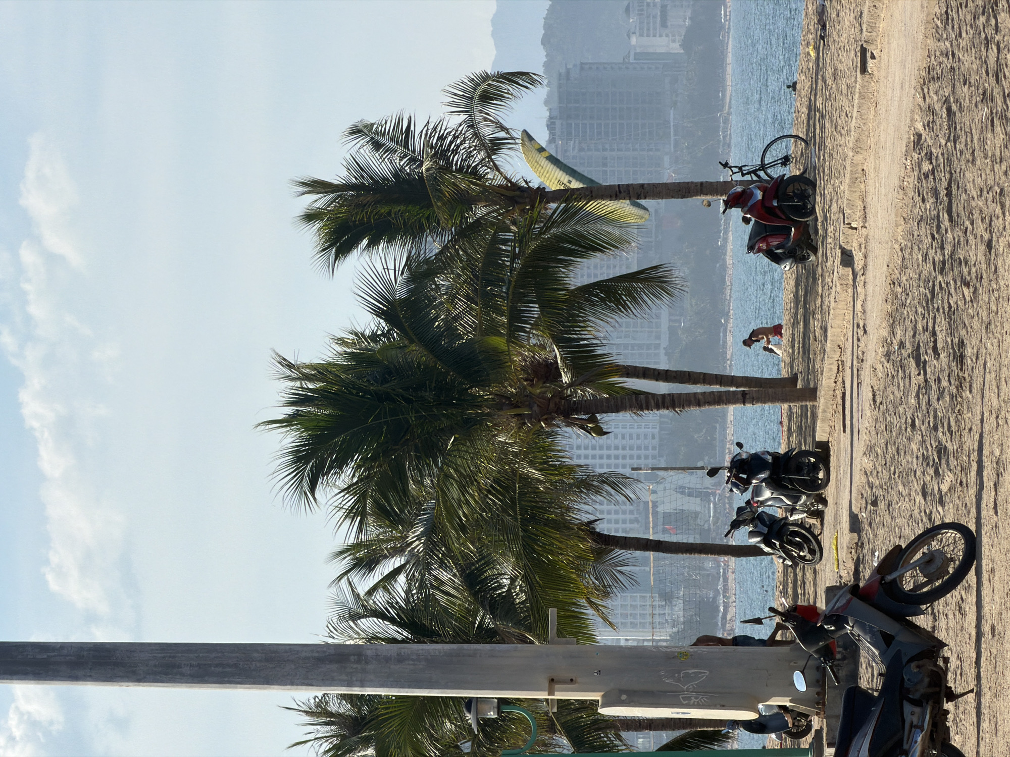 The image captures a section of a beach in Nha Trang Ward, Vietnam. A large, grey concrete pole stands prominently on the left side of the foreground, with a black lamp fixture attached. Adjacent to the pole, several palm trees with dense green fronds cast shadows on the sandy ground. In the mid-ground, a line of motorcycles is parked on the sand. To the right, a red scooter and a bicycle are parked near the water's edge. Several individuals are visible on the beach and in the water in the distance. The background features a hazy cityscape with numerous buildings and a body of water, likely the ocean. The sky is a pale blue with scattered white clouds. A yellow and green paraglider is visible attached to one of the palm trees.