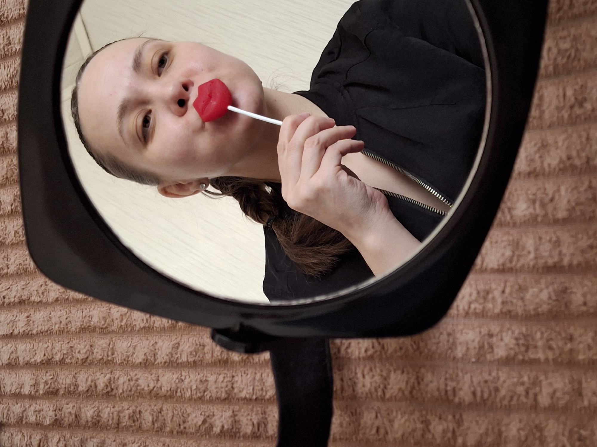 A person is reflected in a black-framed mirror. The person, with hair braided to the side, holds a red, lip-shaped lollipop in their mouth. They are wearing a black jacket with a visible zipper. The background, partially visible around the mirror, consists of a light brown, textured wall. No specific location or identifying details about Chaikovsky, Russia, are visible in the image.