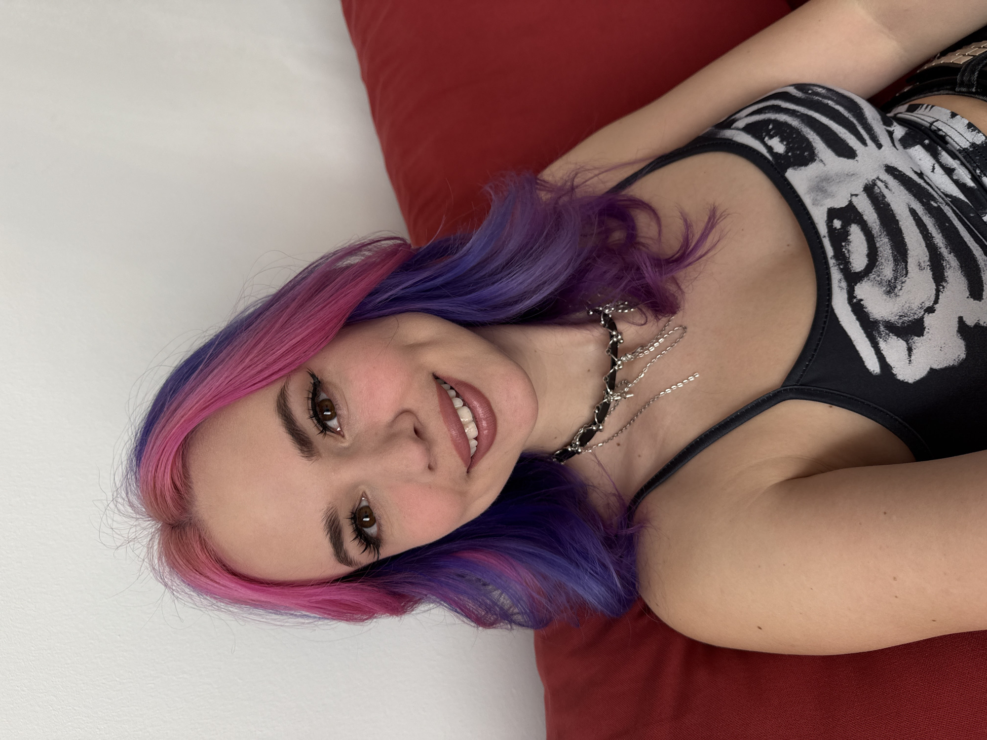 A young woman lies on a red cushion against a white wall, looking at the camera and smiling. She has pink and purple dyed hair and is wearing a black tank top with a white design. She has a layered silver necklace.