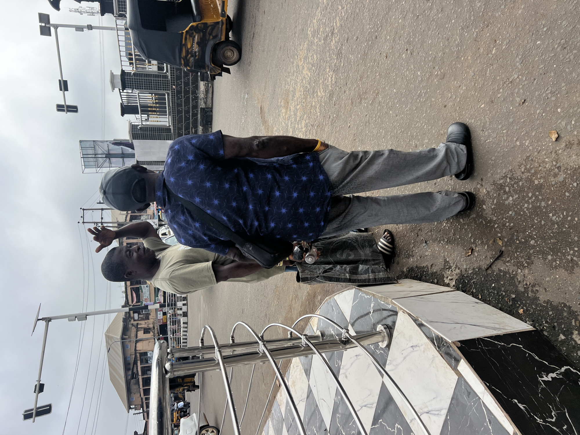 Two men are standing on a paved street in Nkpor, Nigeria, under an overcast sky. The man on the left, wearing a light green t-shirt, is gesturing with his right hand, as if speaking. He has dark skin and short, dark hair. He is wearing dark-striped sandals. Next to him, a man wearing a dark blue shirt with a star pattern and light gray pants stands with his back to the camera. He has a black bag slung over his shoulder and a yellow wristband on his left wrist. He is wearing dark, clog-like shoes. A large, dark gray plastic bag rests on the ground at the feet of the man in the green shirt. Behind the men, a yellow and black auto-rickshaw is visible, as are traffic lights, a metal railing with a decorative pattern, and buildings with sloped roofs. The street is wet in places, suggesting recent rain.