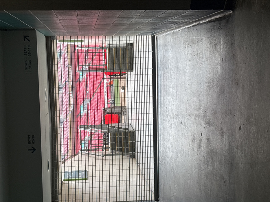 The image presents a view from within a dark, concrete tunnel or concourse, looking out into a brightly lit stadium through a black metal grating. The tunnel features grey concrete walls and a tiled ceiling.
Beyond the vertical metal grate, rows of vibrant red stadium seating are visible, indicating a large arena. On what appears to be the field level, several black structures with red accents are present, resembling stage equipment, large speakers, or temporary event installations. No people are visible, suggesting the image was captured before or after an event, or during the setup/teardown phase. The presence of stage-like equipment hints at a concert or a non-sporting event rather than a typical sports match.
On the left concrete wall of the tunnel, two white rectangular signs with black text are visible. The upper sign reads "ROWS | SEATS" with "37-44 | 95-110" below, accompanied by an arrow pointing right into the stadium. Further down, another sign indicates "ROWS" and "02-30" with an arrow pointing left. A small, partially obscured green sign is situated on the ground level near the bottom left of the tunnel opening, displaying "A&B" and what appears to be a time, possibly "08:56" or "09:56".
The natural light illuminating the stadium area suggests it is daytime. The scene is typical of a large event venue, consistent with the provided location context of Wembley, United Kingdom.