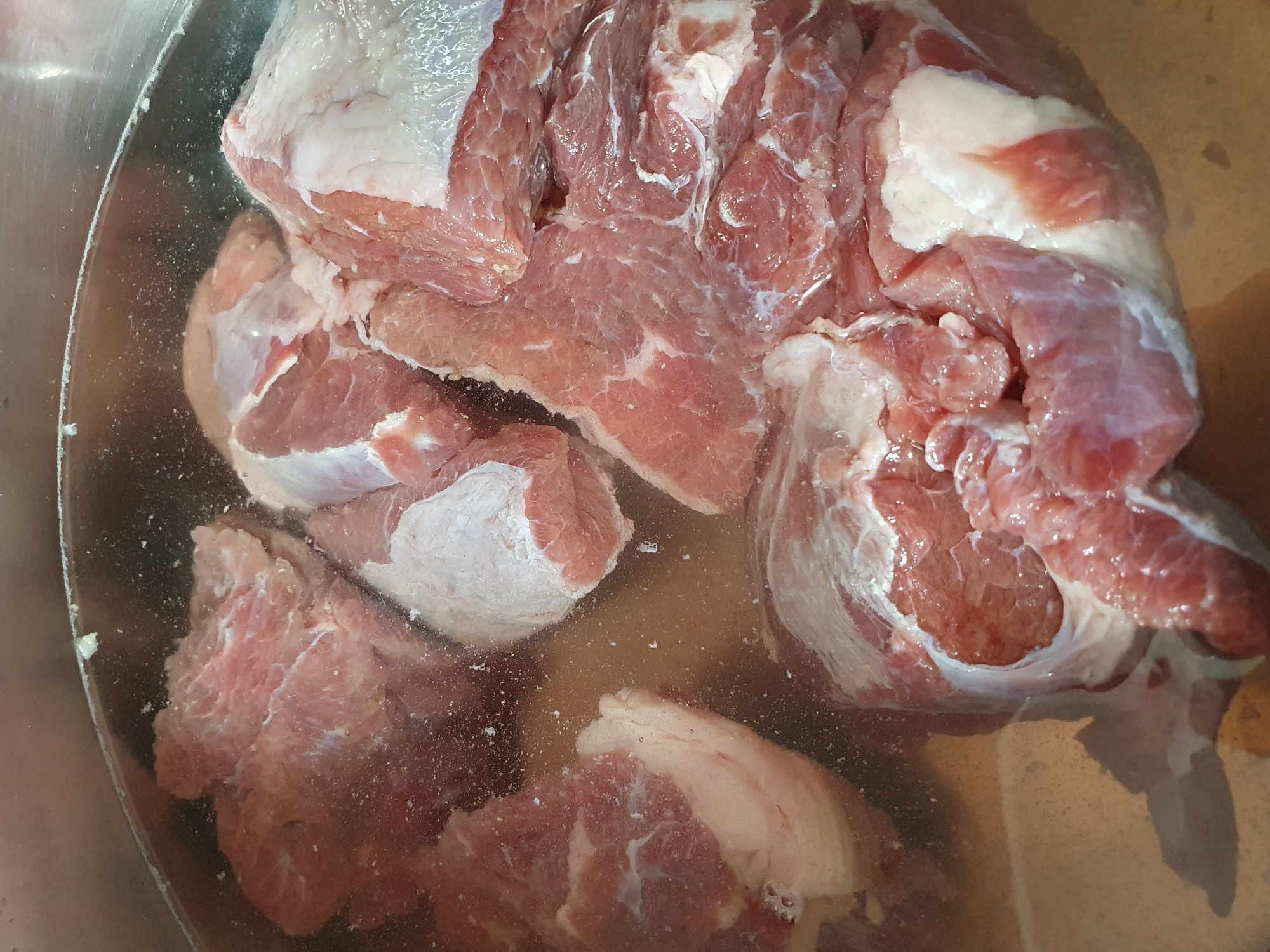A close-up, overhead shot shows several large pieces of raw meat submerged in a clear liquid within a metallic pot. The meat is a reddish-pink color with white streaks of fat. Some pieces have a skin-like covering. Small bubbles are visible in the liquid, and there are white flecks floating on the surface and suspended within the liquid. The pot appears to be made of stainless steel. The lighting is bright, highlighting the texture and moisture of the meat. There is no discernible background or environmental context beyond the pot itself, making it impossible to determine a location in Lusagyugh, Armenia or any other city.