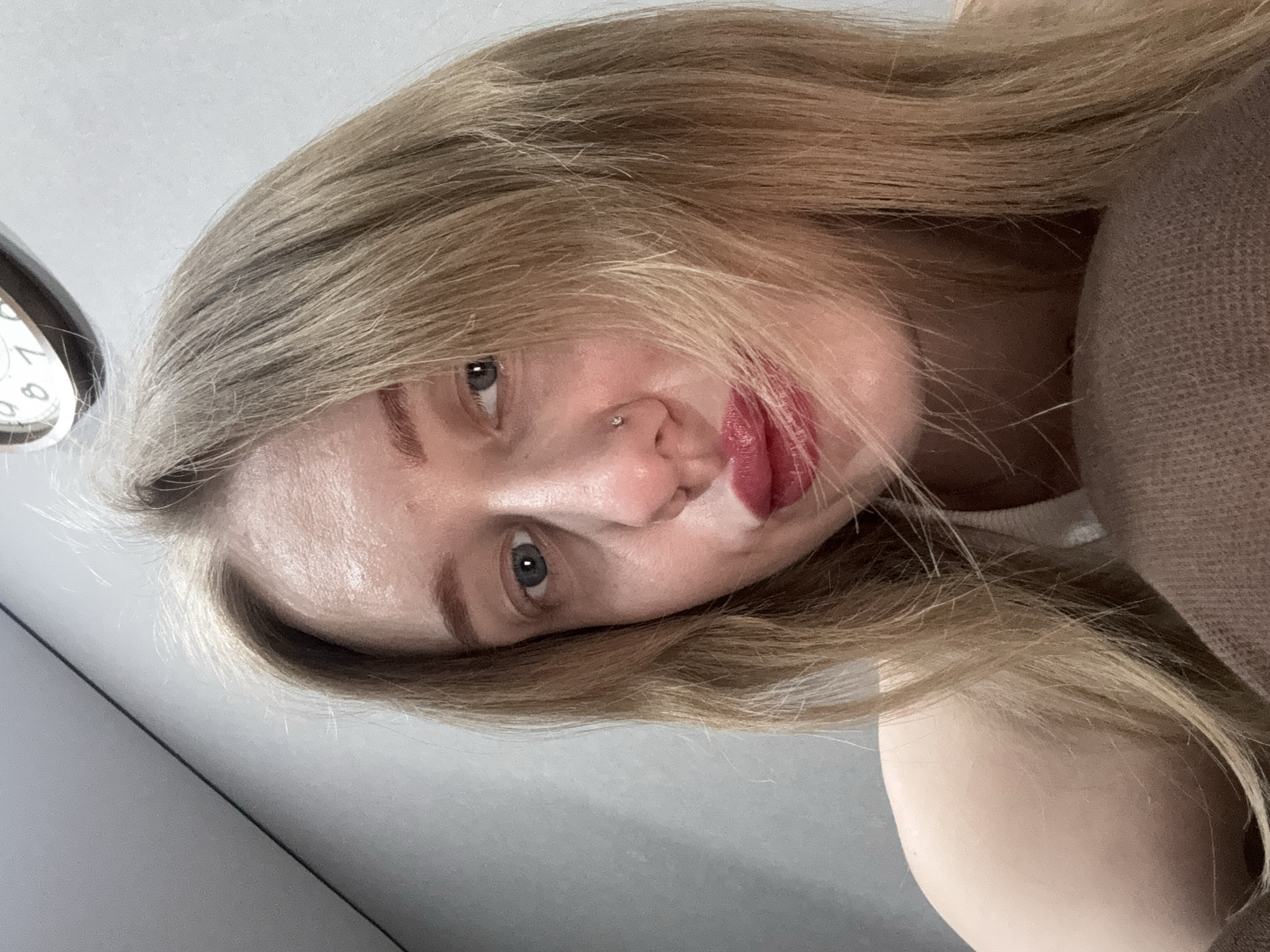 The image displays a close-up, front-facing portrait of a female individual. She has blonde hair that falls around her face and blue irises. A small, silver-colored stud is visible in her left nostril. Her lips are coated with red pigment, and the lower lip appears slightly compressed, with strands of hair adhering to it. A light-colored garment, possibly white, is partially visible on her right shoulder. The lower portion of the frame shows a light brown, textured fabric, consistent with knitwear. The background is a plain light grey wall. In the upper right quadrant, a portion of a round object, consistent with a clock face displaying numerical digits, is visible. The scene is indoors. No specific visual cues in the image confirm its location within Novoaleksandrovka, Russia.