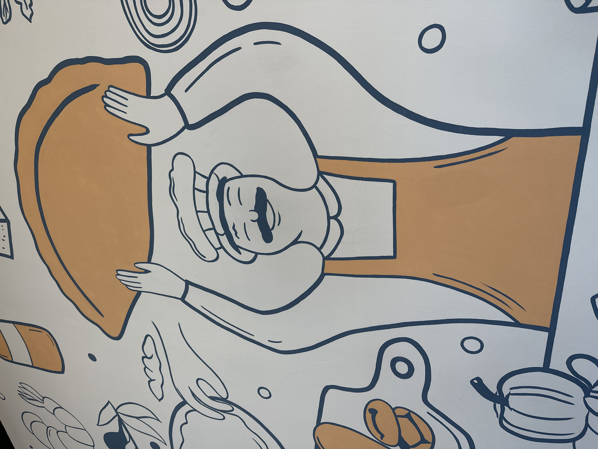 The image depicts a mural on a white wall, likely indoors. The mural features a stylized depiction of a chef in a white chef's hat and apron (colored ochre). The chef appears to be stretching or working on a large rectangular piece of dough, also colored ochre. The chef has hands on either side of the dough, appearing to be in the process of shaping it. Around the chef and the central figure are other food-related items and shapes, also depicted in the same stylized, outlined manner. This includes concentric circles, a cutting board with oval shapes on it, leaves, what looks like a shrimp, and other abstract shapes. The lines are mainly dark blue. The location mentioned, Votkinsk, Russia, cannot be directly verified from the image alone.