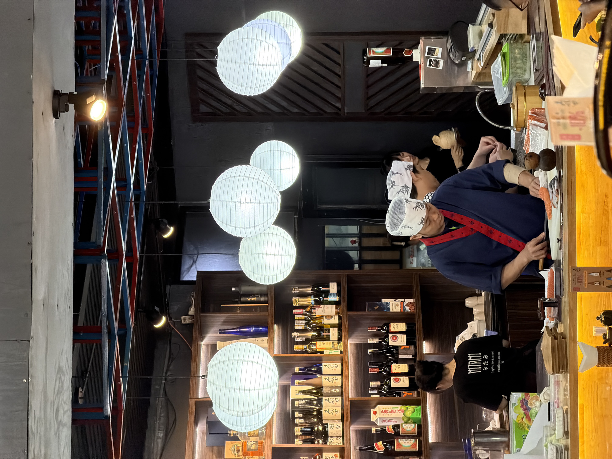 The image displays the interior of a Japanese restaurant, likely a sushi bar, during what appears to be evening hours due to the artificial lighting. A chef, wearing a blue uniform with a red sash and a white patterned head covering, is the central figure, diligently preparing food, specifically slicing a piece of salmon, on a wooden cutting board behind a counter. Another staff member, dressed in a black t-shirt, is partially visible behind the counter to the chef's left, also engaged in work. Customers, some wearing white patterned head coverings similar to the chef's, are seated on the opposite side of the counter, observing the food preparation.
The restaurant's decor features several large, glowing white spherical paper lanterns suspended from the ceiling, casting a soft light. Behind the counter, extensive dark wooden shelves are filled with a diverse selection of bottles, including wines, sake, and other spirits, illuminated by warm spotlights. A distinctive red and blue lattice-like structure is visible on the ceiling or an upper wall section. The counter surface is made of light-toned wood.
Visible text includes "NITRATI" (or similar stylized font), "Japan Restaurant", and "Nhatrang" in English, along with Japanese characters, on the black t-shirt of the second worker. Various Japanese script and brand names are also visible on the bottles and packaging on the shelves. The scene captures a focused moment of culinary artistry within a warmly lit, authentic Japanese dining environment.