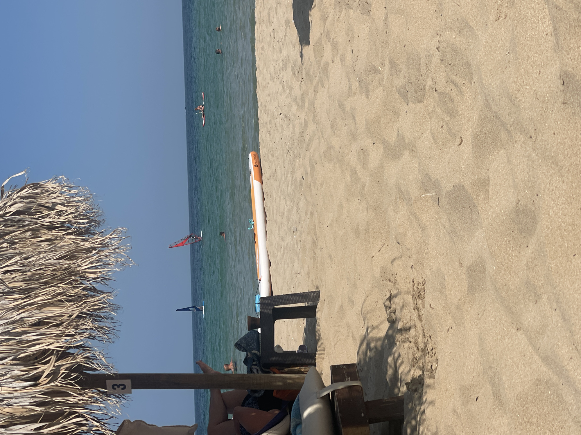 The image depicts a beach scene in Primorsko, Bulgaria, on a sunny day. In the foreground, a thatched umbrella provides shade over a section of sandy beach where a lounge chair is partially visible. To the right of the chair, a small, dark brown woven table holds a bag and a potted plant. A large, orange and white paddleboard lies on the sand near the water's edge.
The sea is a clear turquoise color, with several people engaged in various water activities. Two individuals are windsurfing, one with a red sail and the other with a blue sail. Another person is on a paddleboard further out in the water, and two other people are swimming. The sky is a cloudless, vibrant blue. The overall atmosphere is one of a relaxed summer day at the seaside.