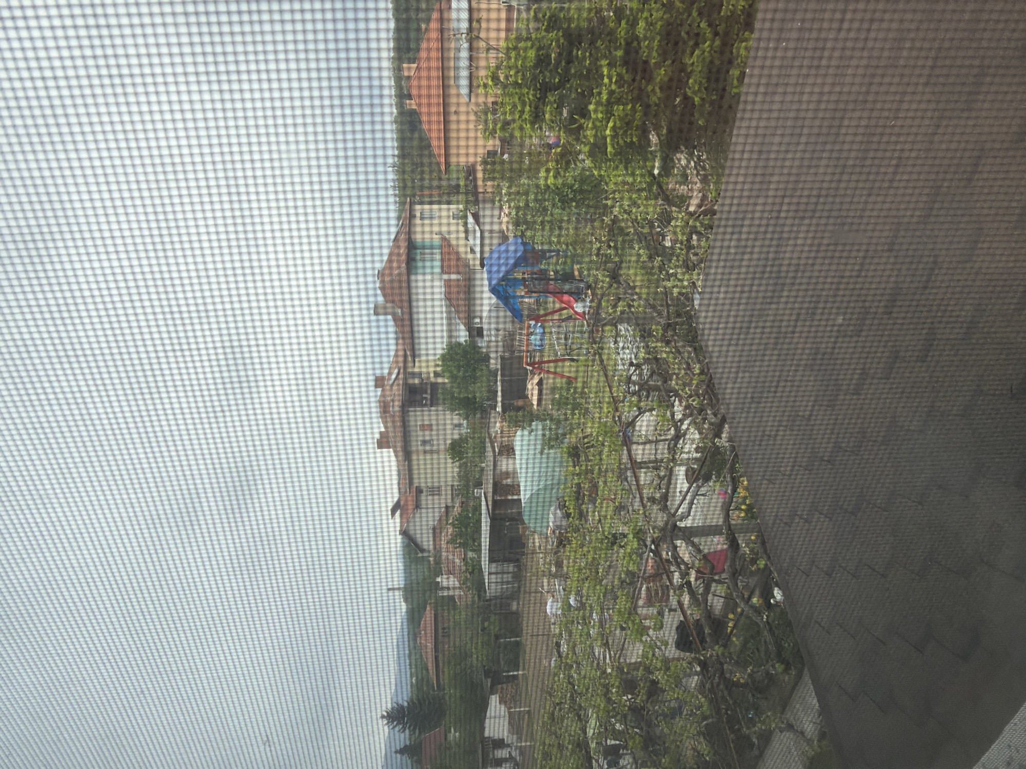 The image, taken from behind a screen, depicts a residential area in Barakovo, Bulgaria. Several houses with red-tiled roofs are visible in the background. A children's playground with blue and red play structures is located in the foreground amongst trees and greenery. There are also some small sheds and tents among the vegetation. The image has an obstructed view due to the screen mesh.