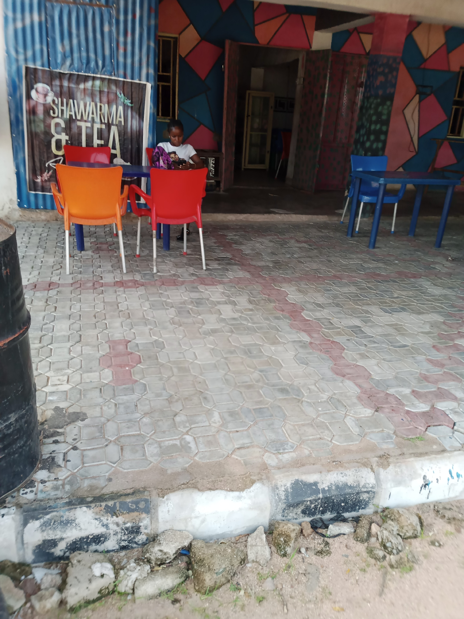 An outdoor scene in Jalingo, Nigeria, depicts the entrance to a shop with colorful geometric patterns on the walls. In front of the shop, several plastic chairs of orange and red hues surround blue tables on a cobblestone pavement. A young person is seated at one of the tables, engrossed in their mobile phone. Behind the tables, a sign reads "SHAWARMA & TEA," indicating the establishment's offerings. To the left of the scene, a large black cylindrical object, possibly a barrel, stands near the entrance. The surrounding area appears to be a dusty, unpaved ground with scattered rocks and some sparse vegetation.