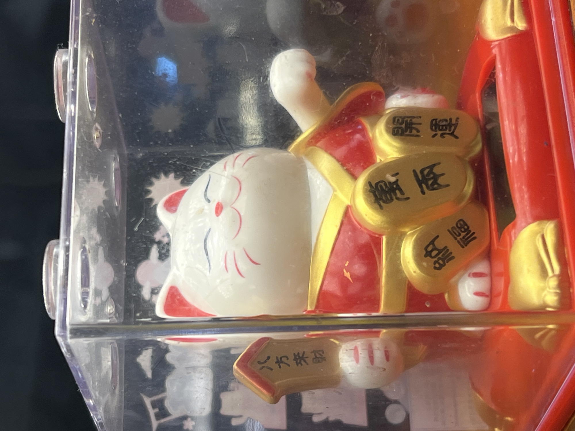 The image shows a close-up of a white Maneki-neko (beckoning cat) figurine housed within a clear plastic box. The cat is wearing a red and gold garment and holding a gold tablet with Chinese characters. There are gold oval shapes with Chinese characters below the cat. The figurine is sitting on a red and gold base. The background is dark and blurred. The scene is likely inside a shop or market stall in Karon, Thailand, given the context provided.