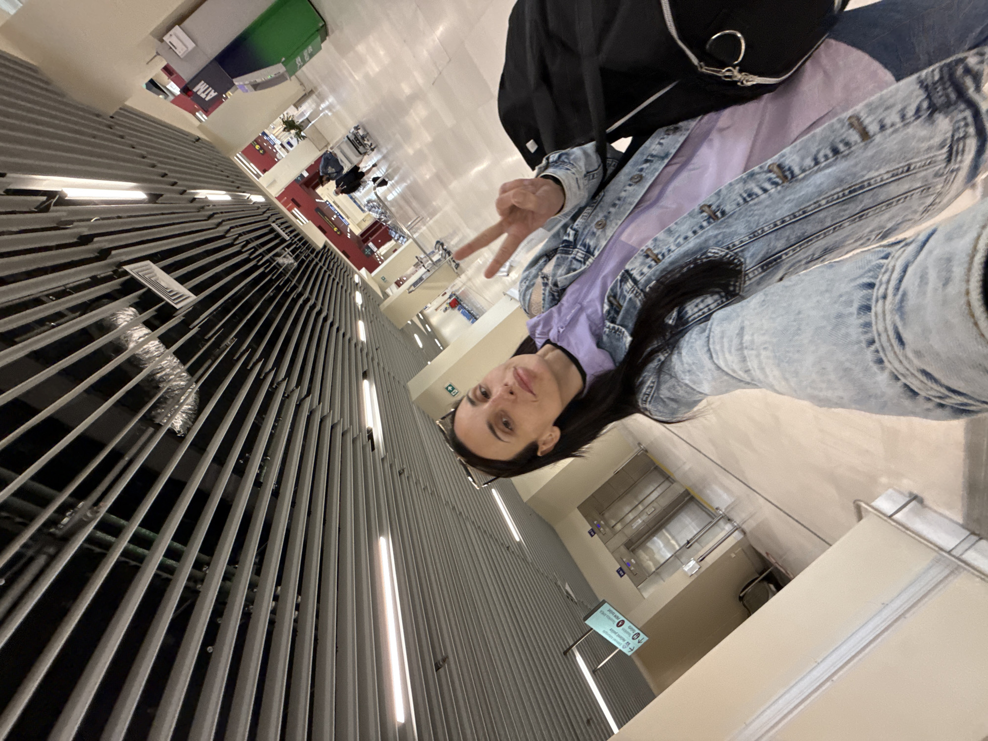 A young woman poses for a selfie indoors at an airport, holding up two fingers in a peace sign. She is wearing a denim jacket, a purple shirt, and a black backpack. The background shows airport infrastructure: seating areas, corridors, signs with symbols and arrows, and ATM machines. The overall setting suggests a travel-related context in Kounoupidiana, Greece, given the airport location.