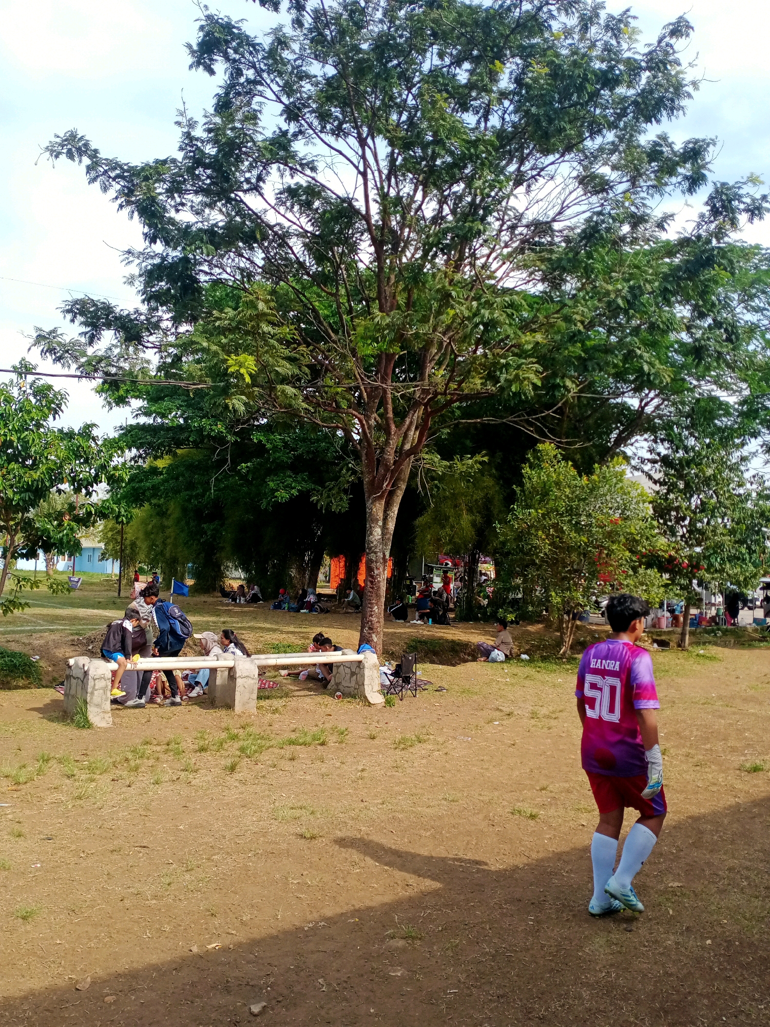 In Cangkuang Wetan, Indonesia, on a dirt field beneath a large tree, a young person wearing a purple and pink soccer jersey with the name "HANORA" and the number "50" on the back, stands facing away from the camera. The individual is also wearing red shorts, white socks, and soccer cleats. Other people are visible in the background, some sitting on a stone bench, while others are gathered on blankets or the ground under the shade of trees. A blue flag is planted in the ground near the bench. The environment is predominantly earthy with scattered dry grass and some vegetation. The sky is visible in the upper portion of the image, with some clouds.