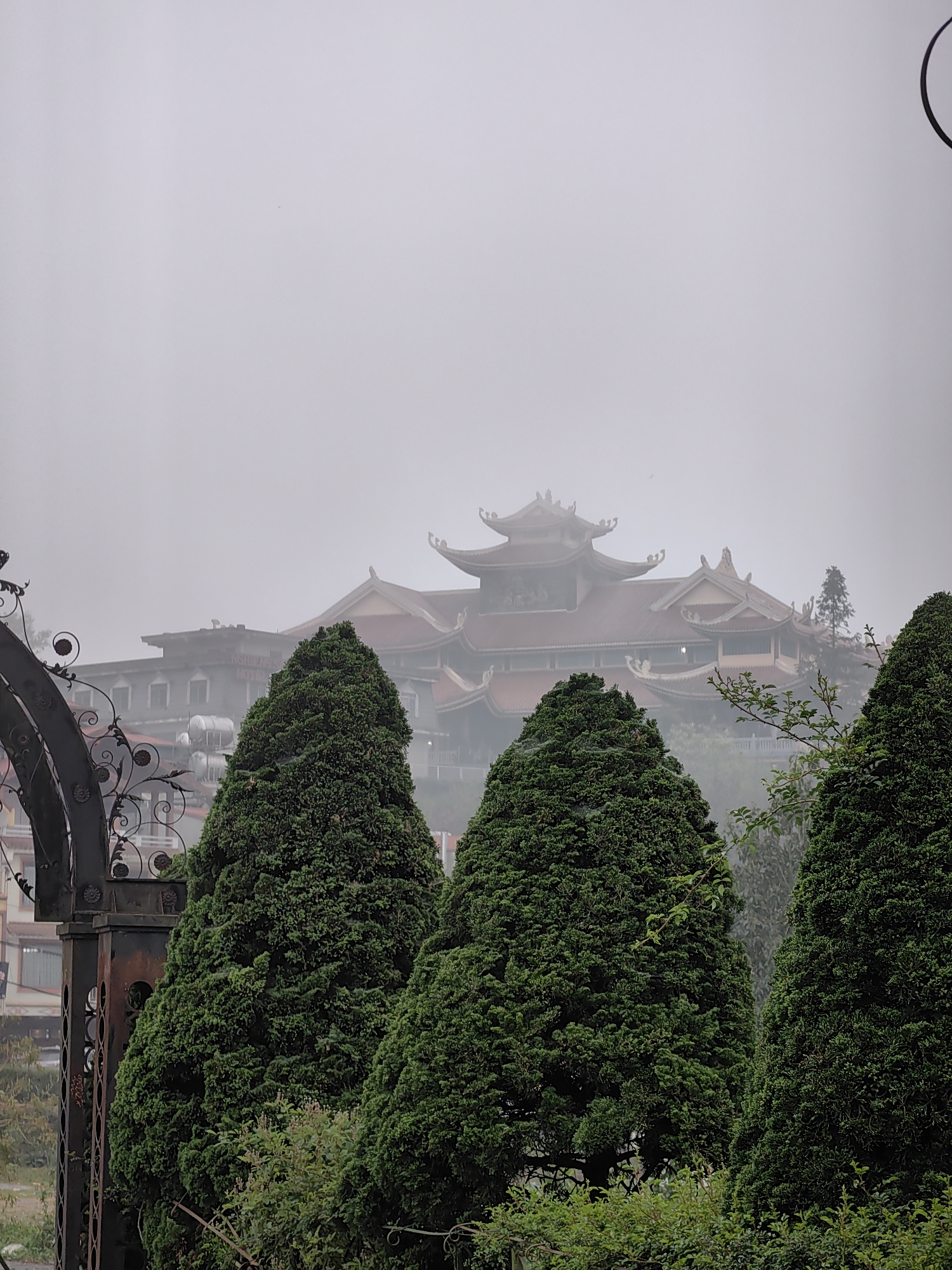 The image presents a panoramic view of an urban landscape in Sa Pa, Vietnam, enveloped in dense, uniform grey mist or fog.
In the immediate foreground on the left, a section of a dark, ornate wrought-iron archway or gate is visible, featuring intricate scrollwork and decorative elements. Across the mid-foreground, several tall, conical evergreen trees with dense, dark green foliage are prominently positioned, partially obscuring the structures behind them. Additional green shrubs and bushes are visible at ground level below these trees.
In the midground, to the left of the central temple structure, a multi-story building, identified as "NGHIÊN S HOTEL" from its partially legible rooftop sign, is visible through the fog. This hotel building features multiple windows and what appear to be water tanks on its roof.
Dominating the background and situated on an elevated position is the Van Hanh Pagoda, a large temple complex. Its architecture is characterized by multi-tiered, reddish-brown tiled roofs with distinctive curved eaves and ornate ridge decorations, typical of traditional Vietnamese temple design. The complex comprises several connected structures and levels. The pervasive fog diffuses light and softens the details of all distant structures. No people or discernible actions are visible in the static scene.