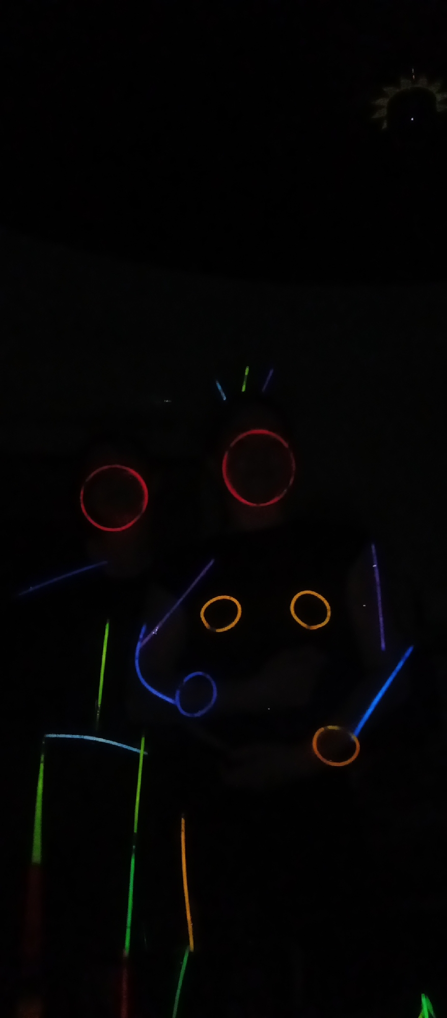 The image shows a dark room with several glowing objects. There are three large circles that are red or orange in color. There are also several smaller circles, some of which are blue and some are orange. Additionally, there are several lines that are various colors, including green, blue, and orange. In the top right corner of the image, there is a bright light that appears to be a sun-shaped decoration. The image is grainy and dark, but the glowing objects are clearly visible.