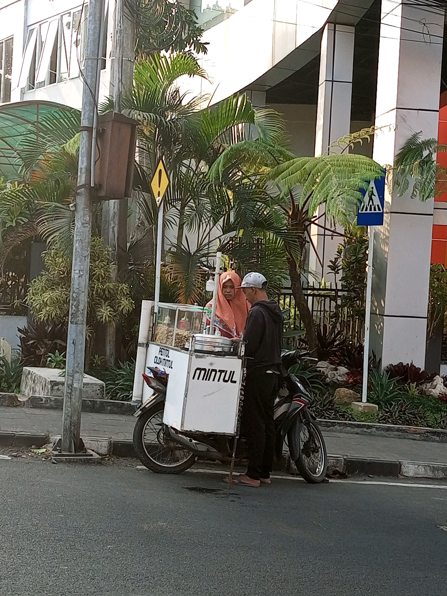 In Batu, Indonesia, a street vendor operates a mobile food cart, likely selling "pentol" (a type of Indonesian meatball). Two individuals are present near the cart. The vendor, a woman wearing an orange hijab, stands behind the cart. To her right, a man in a dark hoodie and a light-colored cap is facing her, suggesting an interaction. The cart itself is white with red lettering that reads "PENTOL OLUL MINTUL" and "MINTUL." It's attached to a motorcycle, indicating its mobile nature. The scene is set on a paved street next to a sidewalk lined with lush tropical vegetation, including palm trees and ferns. In the background, a modern building with white and grey accents is visible, along with a pedestrian crossing sign. A lamppost with a brown electrical box stands prominently to the left of the frame. The lighting suggests it is daytime, with the sun casting shadows.