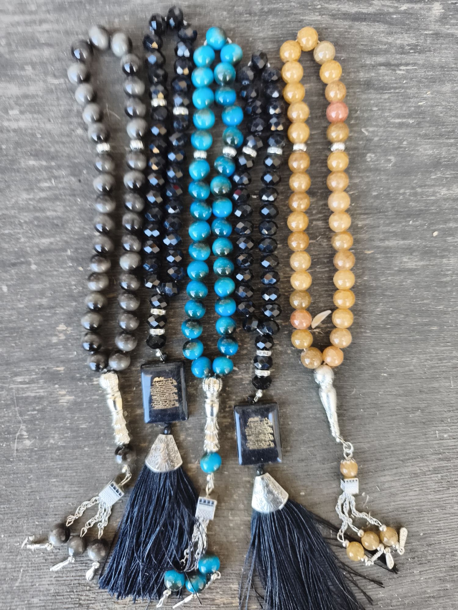 A top-down, medium shot captures four strands of prayer beads, also known as tasbih, laid out on a weathered wooden surface. The beads vary in color and material, with some appearing to be polished stone and others faceted crystal. From left to right, the first strand features gray, opaque beads. The second strand is composed of black, faceted crystal beads. The third strand is made of vibrant blue, rounded beads. The fourth strand consists of translucent, amber-colored beads, with one bead exhibiting a peachy hue. Each strand is adorned with decorative silver findings and tassels. The two leftmost strands have a dark, rectangular pendant with Arabic calligraphy. The two rightmost strands have silver conical caps and tassels. The wooden surface displays a textured grain and appears to be a tabletop or a floor. There are no people visible in the image. The lighting suggests an outdoor or well-lit indoor environment. The image is a static, object-focused composition with no discernible narrative or action. The scene is devoid of any specific geographical markers that would indicate a location in Siazan, Azerbaijan.