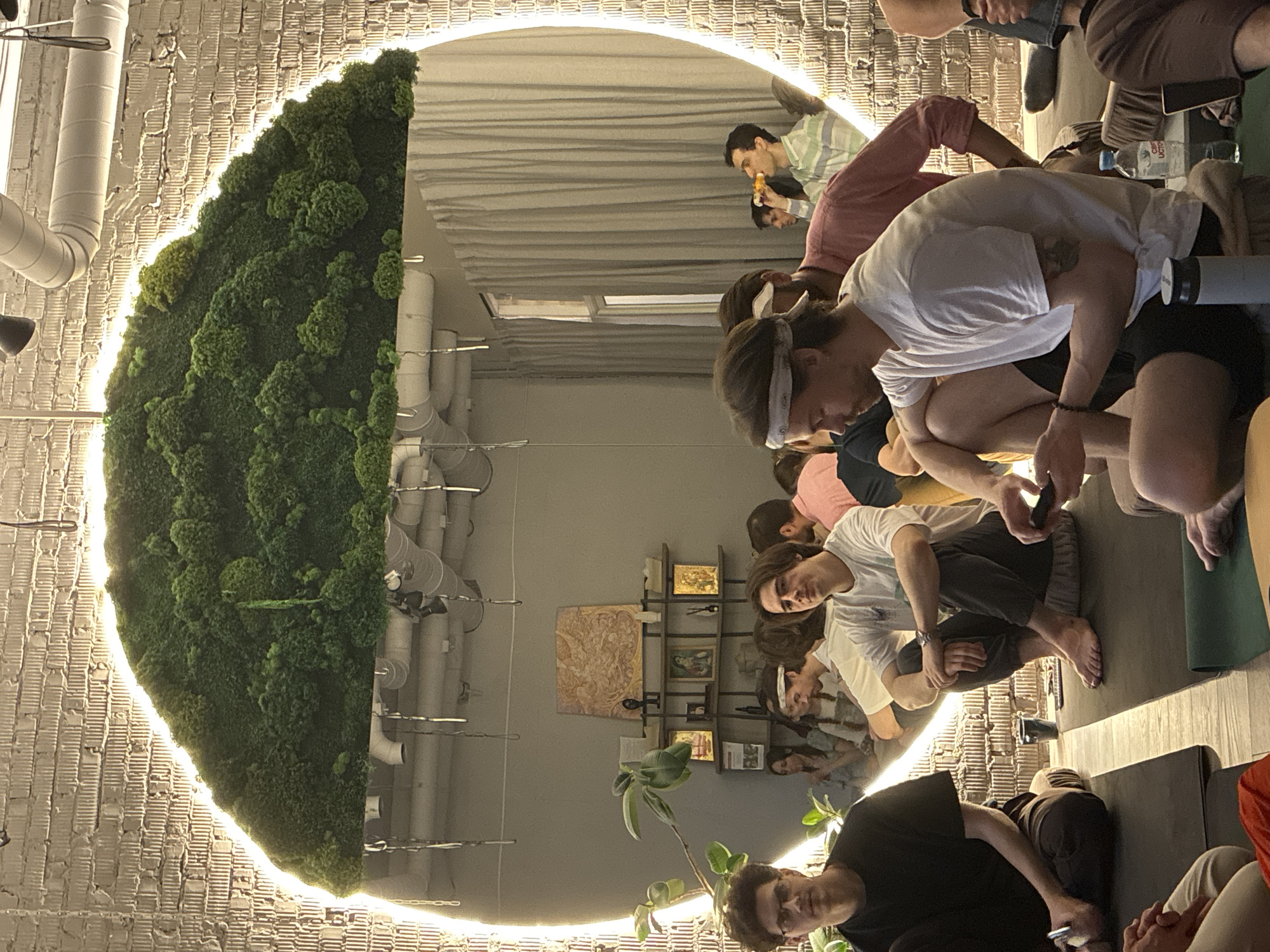 A brightly lit, semi-circular room in what appears to be Moscow, Russia features a living plant wall on one side and a large mirror on the other, reflecting the scene. Around a yoga studio, there are around 10 people in yoga poses. The lighting is bright, casting an even glow. One man is eating a slice of pizza. Shelves with art are on the far wall.