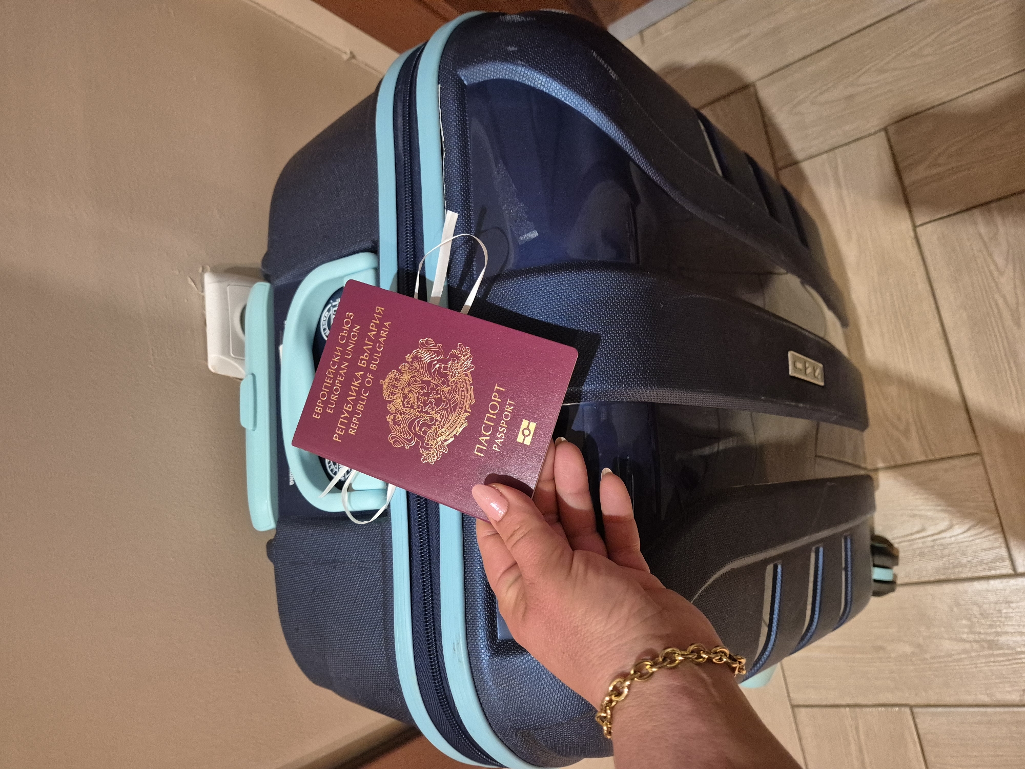 A person's hand, adorned with a gold bracelet and a pale pink manicure, holds a red Bulgarian passport in front of a dark blue and teal suitcase. The suitcase has visible zippers and straps. The background is a neutral-colored wall and a light-colored tiled floor. An electrical outlet is visible on the wall behind the suitcase.