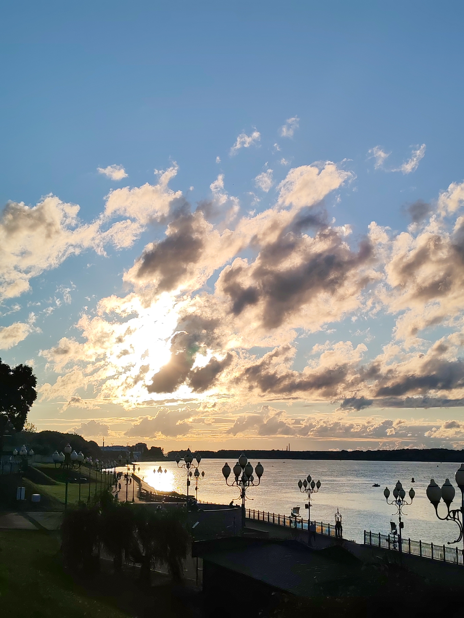 This is a vertical, eye-level, full shot of a waterfront promenade at sunset. The sky is a vibrant blue with scattered, fluffy clouds catching the golden light of the setting sun. The sun itself is partially obscured by clouds on the left side of the frame, casting a bright, shimmering reflection on the wide body of water.
Along the water's edge, a dark promenade is visible, lined with ornate lampposts. People are walking along the promenade, silhouetted against the water. A grassy embankment slopes down to the promenade on the left side of the image, dotted with more lampposts and some trees. In the distance, the opposite bank of the water is lined with trees and some distant structures. Several small boats are scattered across the water. The overall mood is peaceful and serene, capturing the beauty of a sunset over a city waterfront.