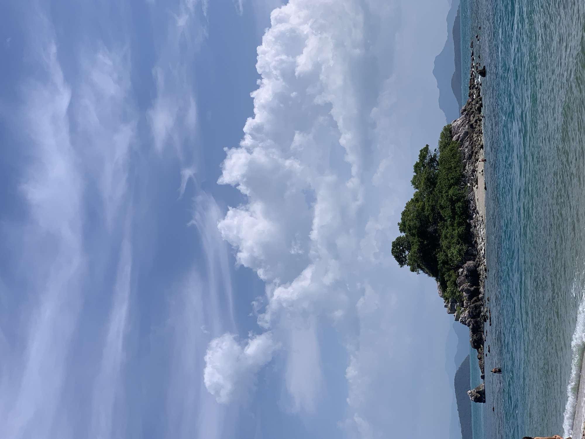 The image depicts a tropical island with lush green vegetation and rocky shores, surrounded by clear blue water. In the foreground, a sandy beach with gentle waves is visible. Further out, a small island with trees and rocks stands in the ocean. Several people are in the water, appearing to swim or wade. In the background, more islands and mountains can be seen under a partly cloudy sky. The scene conveys a sense of tranquility and natural beauty. The location is likely a coastal area in Thailand.