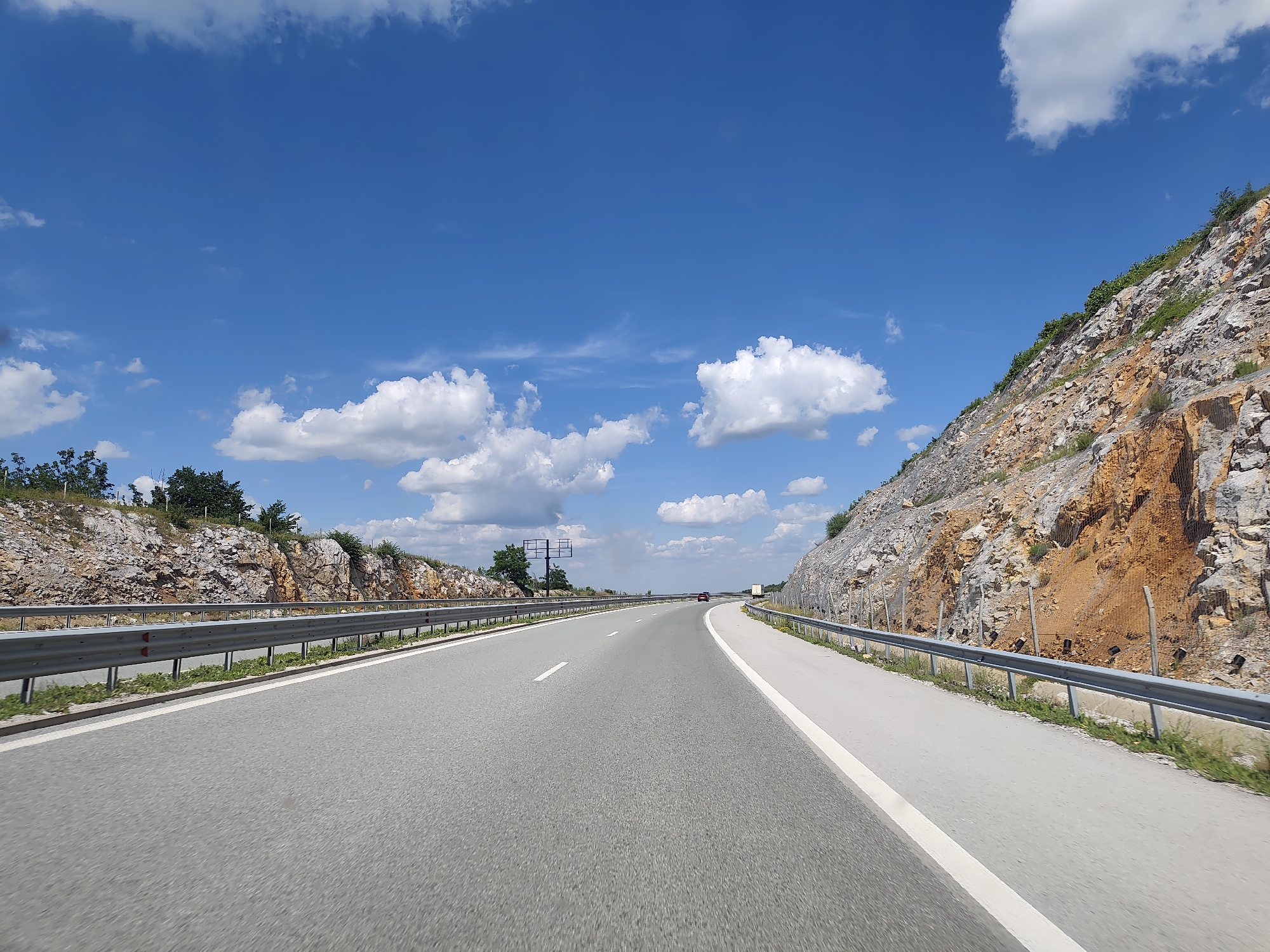 The media file depicts a clear, sunny day with a bright blue sky dotted with fluffy white clouds. The scene is a highway cutting through a mountainous landscape. The road itself is asphalt, with white lane markings and a guardrail running along the right side. On either side of the highway, rocky hills rise steeply, with some vegetation growing on their slopes. The rocks display a mix of grays, browns, and orange hues.
The perspective suggests the image was taken from a moving vehicle, looking forward down the highway. The activity taking place is travel on the highway. A few distant vehicles are visible on the road.
Notable details include the time of day, which appears to be midday due to the strong sunlight and the shadows cast. The weather is fair and sunny. The landscape, with its rocky formations and the highway infrastructure, provides location cues that are consistent with a mountainous region. There is no visible text in the image. The overall mood is one of open road travel and natural scenery.