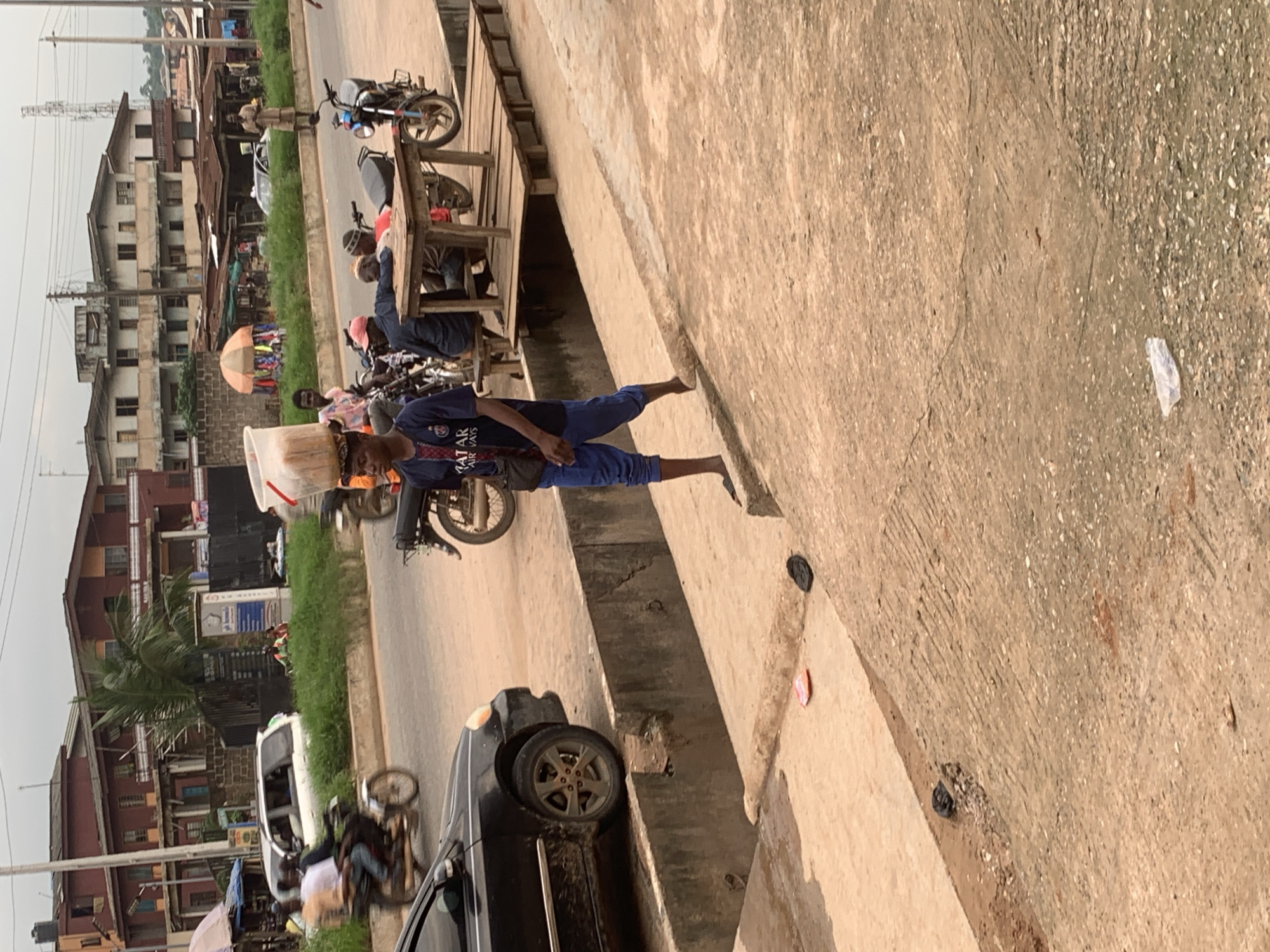 This is a full-frame, eye-level, outdoor, daytime photograph taken on a street in Shagamu, Nigeria. The foreground is dominated by a rough, dusty concrete surface with some small pebbles and debris. A slight incline leads towards the middle ground. A narrow, concrete drainage ditch runs along the left side of the foreground, with a light-colored object, possibly a piece of paper or plastic, resting on its edge. Further down the ditch, a black sandal is visible on the concrete.
In the middle ground, a young man, appearing to be in his late teens or early twenties, is walking on the concrete pathway. He is wearing a dark blue t-shirt with white lettering and blue pants. A dark bag is slung across his chest. On his head, he balances a large, transparent plastic container filled with what appears to be bread. To his right, the street is visible, with traffic including motorcycles and cars. A black car is parked on the left side of the street. Behind the car, a raised grassy area is present, with more buildings and shops visible in the background. A wooden platform structure with a table and benches is positioned slightly to the right of the walking man.
The background depicts a bustling urban environment with several buildings, some appearing to be residential or commercial structures with multiple stories. A few people are visible on motorcycles and near shops. The sky is overcast, suggesting a cloudy or hazy day. The overall scene captures a slice of daily life in a Nigerian city, highlighting street commerce and transportation.
