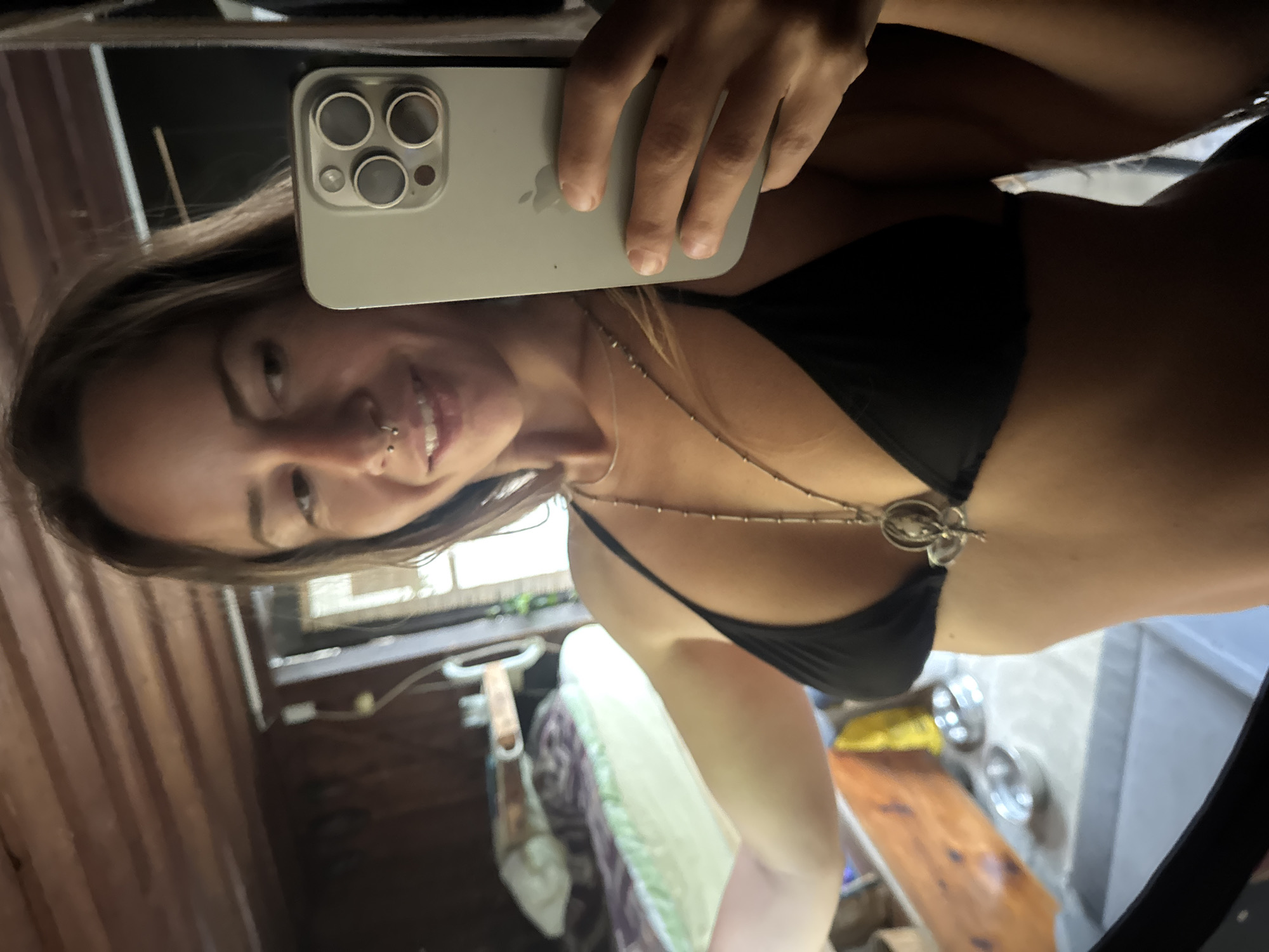 A woman is taking a mirror selfie in a room with wooden walls and ceiling. She is wearing a black bikini top and a necklace with a pendant. She is holding a smartphone in her right hand, and her left arm is extended. In the background, there is a bed with white and purple bedding and some shelves with objects on them.
