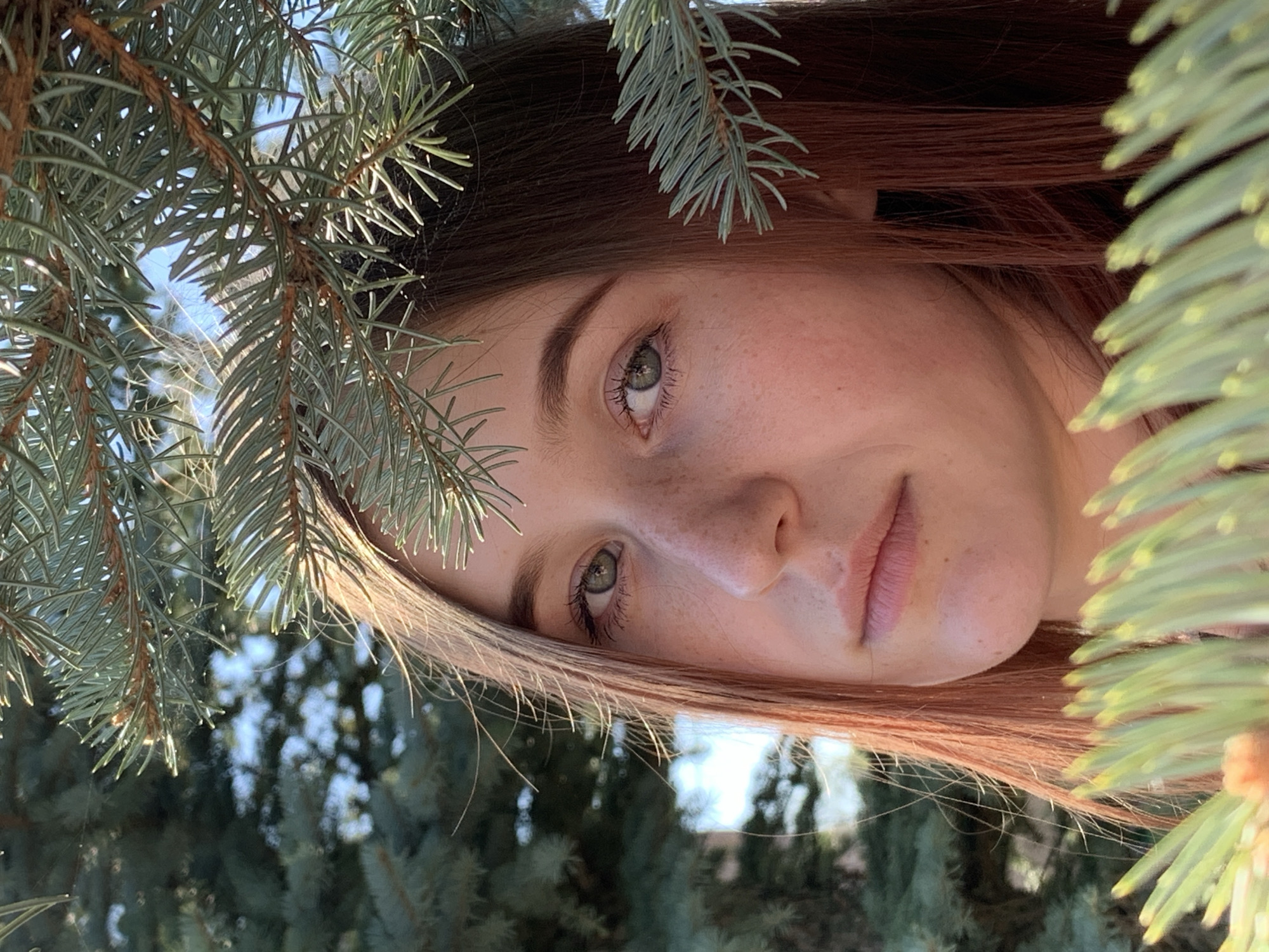 A person with fair skin, freckles, and light green eyes is positioned behind branches of an evergreen tree. The person's hair is reddish-brown, and they are looking directly at the viewer. The background is blurred and appears to be more evergreen trees. The image is rotated slightly, making the person appear to be lying on their side. No information is present in the image to associate it with Фоки, Russia.