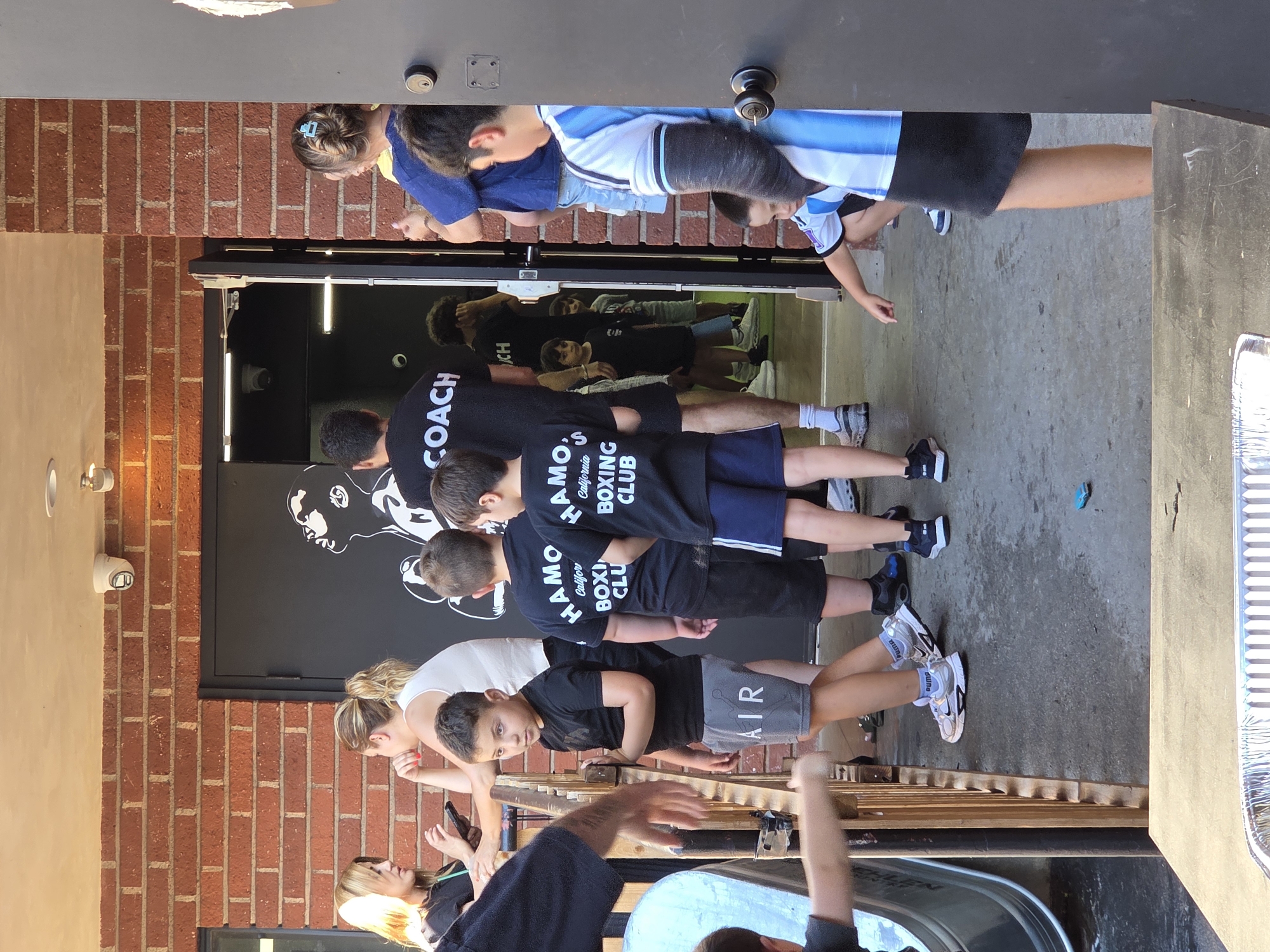 A group of children and adults are standing outside a building with a brick facade and a large black door featuring a graphic of a person looking upwards. Several children are wearing black t-shirts with "HAMO'S CALIFORNIA BOXING CLUB" printed on them, along with athletic shorts or pants. One boy in the foreground wears gray shorts with "AIR" printed on them. Another boy is wearing a blue and white striped shirt and black shorts, with a cast on his left arm. An adult man in the background wears a black t-shirt with "COACH" printed on it. Two women are standing near the entrance, one looking at her phone. The scene appears to be outdoors, with a concrete ground and a wooden railing. The location is identified as Glendale, United States of America.