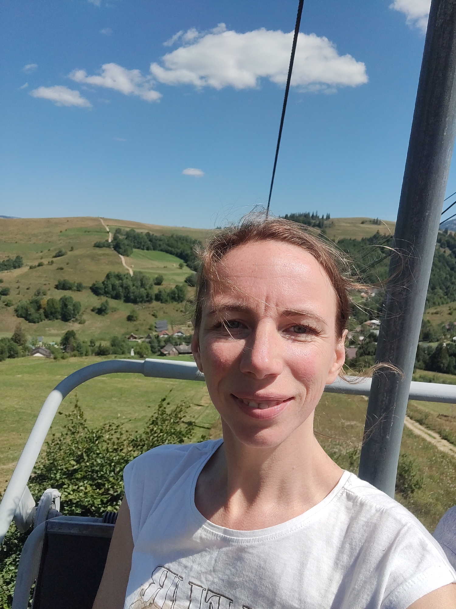 A woman with blonde hair, wearing a white t-shirt, is captured in a selfie. She is seated on a ski lift, with a blurred mountainous landscape in the background. The woman is smiling, and her hair is slightly windblown. The ski lift appears to be in a rural area, possibly the Carpathian Mountains, near the village of Izky, Ukraine. The day is sunny with a clear blue sky and scattered clouds. The ski lift's metal structure is visible in the foreground.