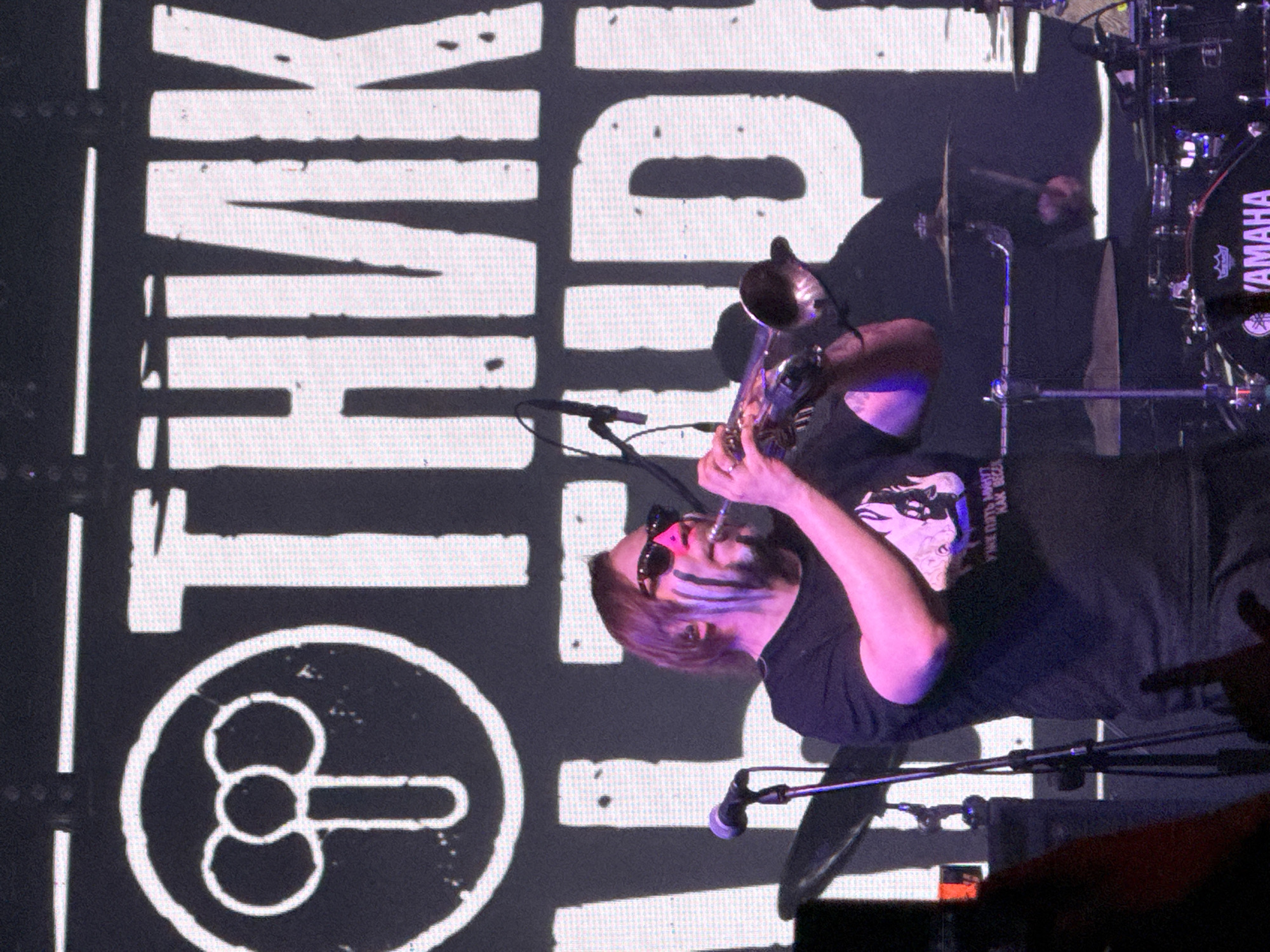 The image depicts a live musical performance on an indoor stage, presumably in Большая Кивара, Russia. In the center, a male musician, estimated to be in his 40s or 50s, is playing a silver-colored trumpet. He has shoulder-length brown hair and a beard, partially covered by vertical black and white face paint on his cheeks. He wears dark sunglasses with a distinct pink nose bridge and a dark, short-sleeved t-shirt with a light-colored graphic. Stage lighting casts a prominent magenta-purple hue on him and his instrument. To his left, a black microphone on a stand is positioned. To his right and slightly behind him, parts of a drum kit are visible, including a black Yamaha bass drum and a cymbal being struck by a drummer's arm. The background is dominated by a large LED screen displaying white Cyrillic text and a graphic on a black field. Partially visible text fragments suggest "ОТНИК" and "АЛЬФОРДИ". To the left, within a white circular outline, a stylized white graphic resembling male genitalia (a phallus with three rounded elements at the top) is prominently displayed. In the bottom-left corner, a partial view of an audience member's head and raised arm is visible.