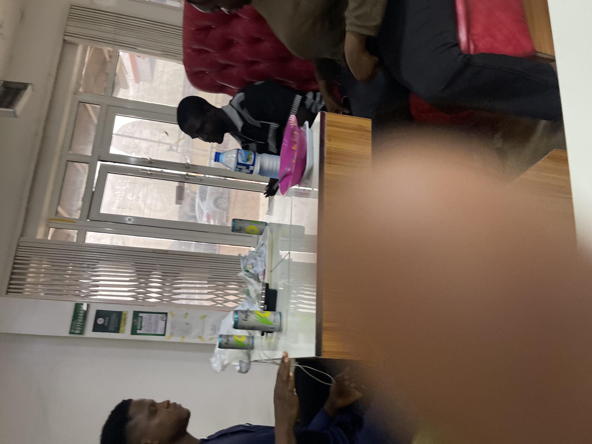 A group of individuals are seated around a table, likely in an office or waiting area, in Jos, Nigeria. The scene appears to be indoors, with a large window in the background featuring a metal accordion gate. The lighting suggests it is daytime.
On the left, a young man wearing a dark blue garment is seated and looking to his right. His hand is visible, resting on the edge of the table.
At the table, several cans of "Fayrouz Pineapple" are placed, along with a white plate holding a pink bowl containing what appears to be food. A bottle of water is also on the table.
To the right of the table, a man in a black and grey striped shirt is seated and looking to his left. Further to the right, a person in a brown top and black pants is seated in a red upholstered chair, their face obscured from view.
The table itself has a light-colored top and a wooden base. In the background, there are some signs on the wall and a glimpse of the outdoor environment through the window, including a car. The overall impression is a casual gathering or meeting.