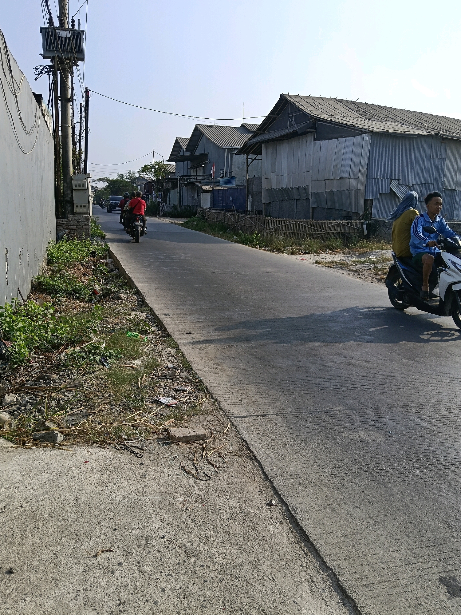 The image depicts a street scene in the city of Belimbing, Indonesia, on a clear day. A paved road runs diagonally from the lower right to the upper left of the frame. On the right side of the road, a white scooter with two occupants is traveling away from the viewer. The driver, wearing a blue jacket and green shorts, is looking to the right. The passenger, a woman wearing a yellow shirt and blue headscarf, is also looking to the right. The scooter is casting a shadow on the road.
To the left of the road, there is a sidewalk or unpaved area with dry vegetation, rocks, and some scattered debris, including a plastic bottle and what appears to be an empty packet. A gray concrete wall runs along the left edge of the image, with electrical equipment mounted on a pole at the far left.
Further down the road, another motorcycle with two occupants is visible, traveling in the same direction as the scooter. The rider is wearing a red shirt, and the passenger's attire is less clear. A black car is following the motorcycle.
On the right side of the road, beyond the scooter, are several buildings with corrugated metal roofs and walls, appearing to be residential structures or small businesses. A wooden fence is visible in front of these buildings. The sky is clear and light blue, indicating sunny weather. The overall environment suggests a modest urban or semi-urban setting.