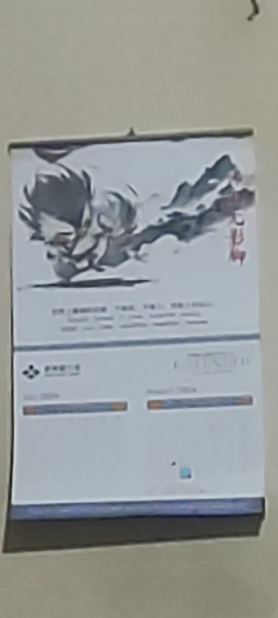 A calendar is hanging on a beige wall. The calendar features a black and white illustration of a person with spiky hair, in a dynamic pose, with a mountainous landscape in the background. Red Chinese characters are visible on the right side of the illustration. Below the illustration, there is text in smaller font, and the bottom portion of the calendar appears to contain a calendar grid with some bars and text. The calendar is attached to the wall at the top. The image is blurry. The location in the city of Gebang, Indonesia cannot be determined from the image.