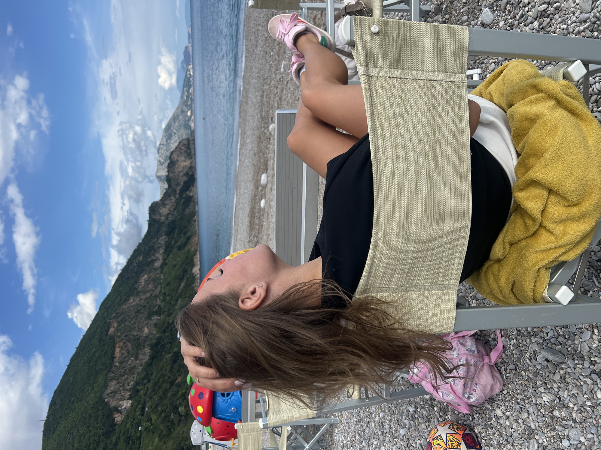 The image captures a young female relaxing on a beige fabric beach chair on a pebble beach in Prijevor, Montenegro, under a bright daytime sky. The subject, likely a child or teenager with long brown hair, is wearing a black top or dress and pink sneakers. She is positioned with her head tilted back, looking upwards, and her hands are near her head, suggesting a moment of relaxation or contemplation. Her legs are crossed and propped up on the chair's frame.
Beside her on the chair is a fluffy yellow towel. On the gravel ground, a small pink backpack adorned with unicorn designs is visible, along with a partially seen colorful soccer ball. A red, blue, and yellow ladybug-shaped toy or container rests near her head.
The setting is a picturesque coastal or lakeside environment. The foreground is a coarse pebble beach, which transitions into a calm body of water with gentle ripples. In the background, majestic, green-forested mountains rise steeply from the water's edge, stretching across the horizon. The sky is blue with scattered white clouds, indicating clear and sunny weather. The overall scene conveys a sense of tranquility and leisure in a natural, scenic location.
