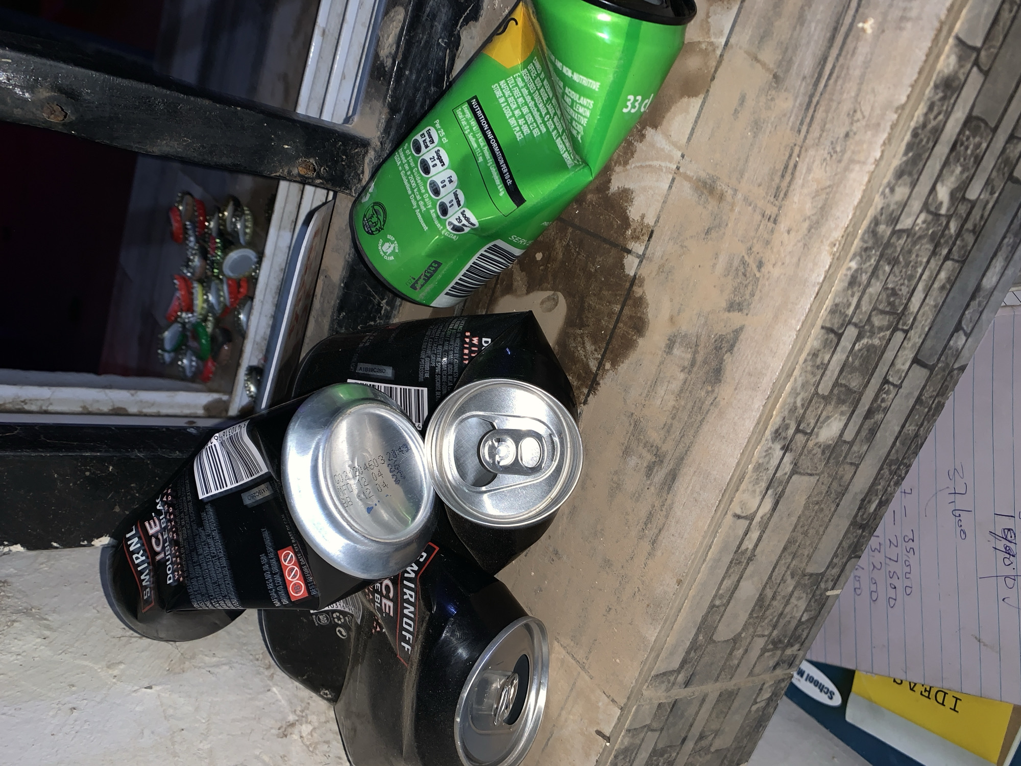 The scene depicts a collection of discarded beverage cans, including "Smirnoff Ice Double Black" and a green Schweppes can, arranged on a textured surface resembling stone or brickwork. A handwritten note with numerical calculations is also present. The cans are crushed or dented, suggesting they are post-consumption waste. The "Smirnoff Ice" cans are primarily black with silver tops, and the Schweppes can is green with yellow accents. The background features a glimpse of what appears to be metal bars and other refuse, possibly bottle caps. The context of New Stadium, Nigeria, is not explicitly evident from the visual data.