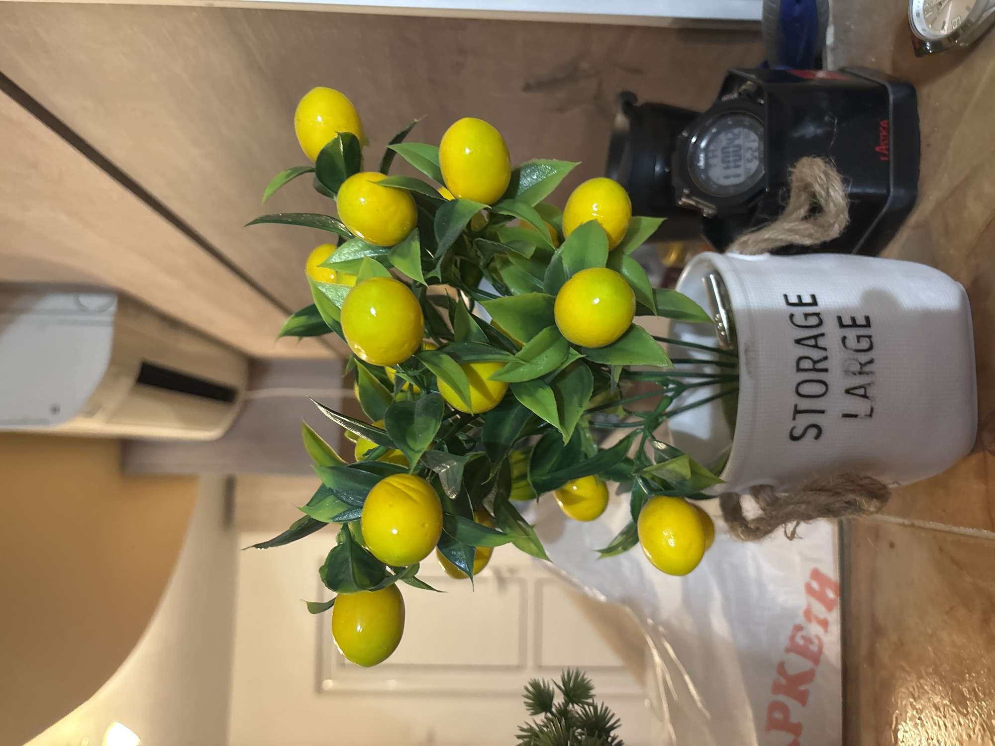 A close-up, slightly high-angle shot depicts a decorative arrangement of artificial lemons and greenery in a white pot. The pot has the words "STORAGE LARGE" printed vertically on its side, and a rope tie is around its upper portion. Behind the plant, there is a black digital watch with a rope tie and a partially visible white watch. The background includes a wall with wood paneling and a section of an air conditioning unit on the left. A shopping bag is partially visible beneath the plant pot. The image was most likely taken indoors. The location in the city of Asenovgrad, Bulgaria, is not discernible from the image. No people are visible in the scene.