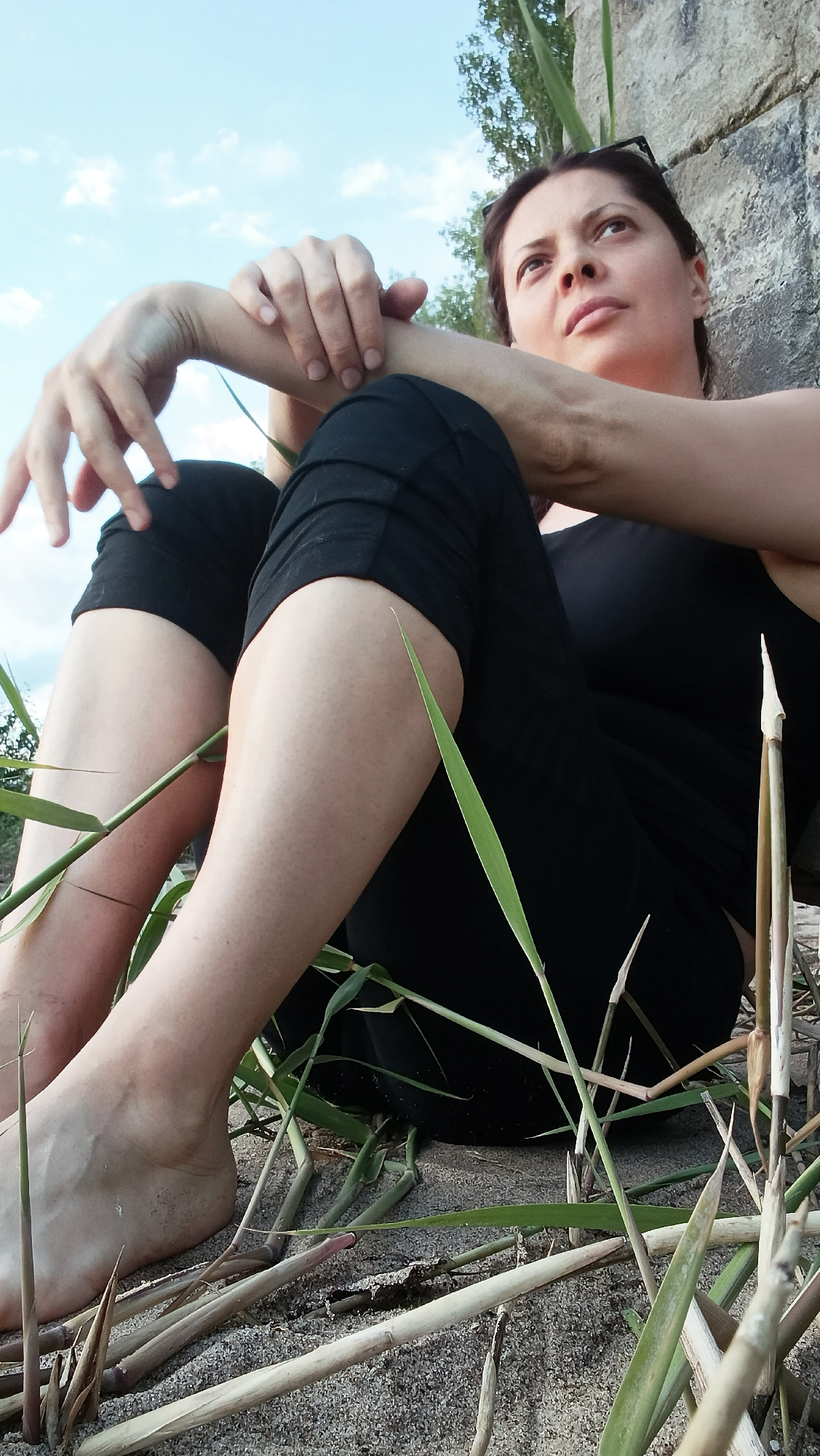 A woman is seated on sand, leaning against a rough concrete structure. She is wearing black leggings and a black tank top. Her arms are crossed, and her gaze is directed upwards and to the right. Green grass and dry reeds are in the foreground. The sky is visible above, with some scattered clouds. The scene appears to be outdoors, possibly near a body of water, given the sandy ground and reeds. The woman's position suggests a moment of reflection or contemplation. The overall lighting indicates it is daytime, likely late afternoon or early morning due to the angle of the sun.