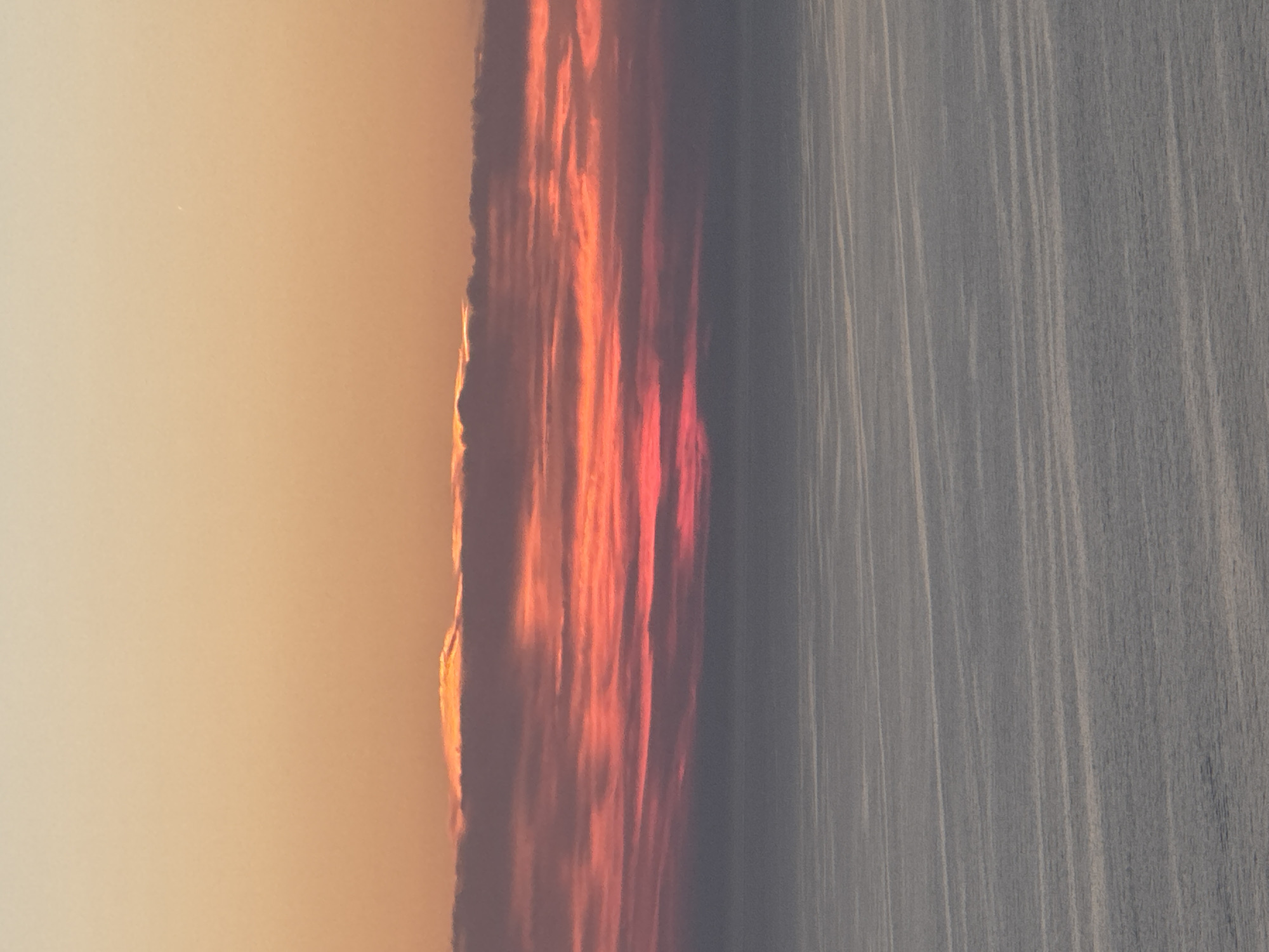 This is a photograph of a sunset over the ocean, possibly from the coast of Zagora, Montenegro. The sky transitions from a pale orange at the top to darker shades of pink and red as it nears the horizon. A band of clouds, illuminated by the setting sun, runs across the middle of the frame, with the lower edge of these clouds appearing a muted gray. Below the clouds, the ocean stretches to the horizon, reflecting the sky's colors in muted tones of gray and blue, with subtle lines of light suggesting ripples on the water's surface. The scene is tranquil and devoid of any human presence or man-made structures. The focus is entirely on the natural spectacle of the sunset and the sea.