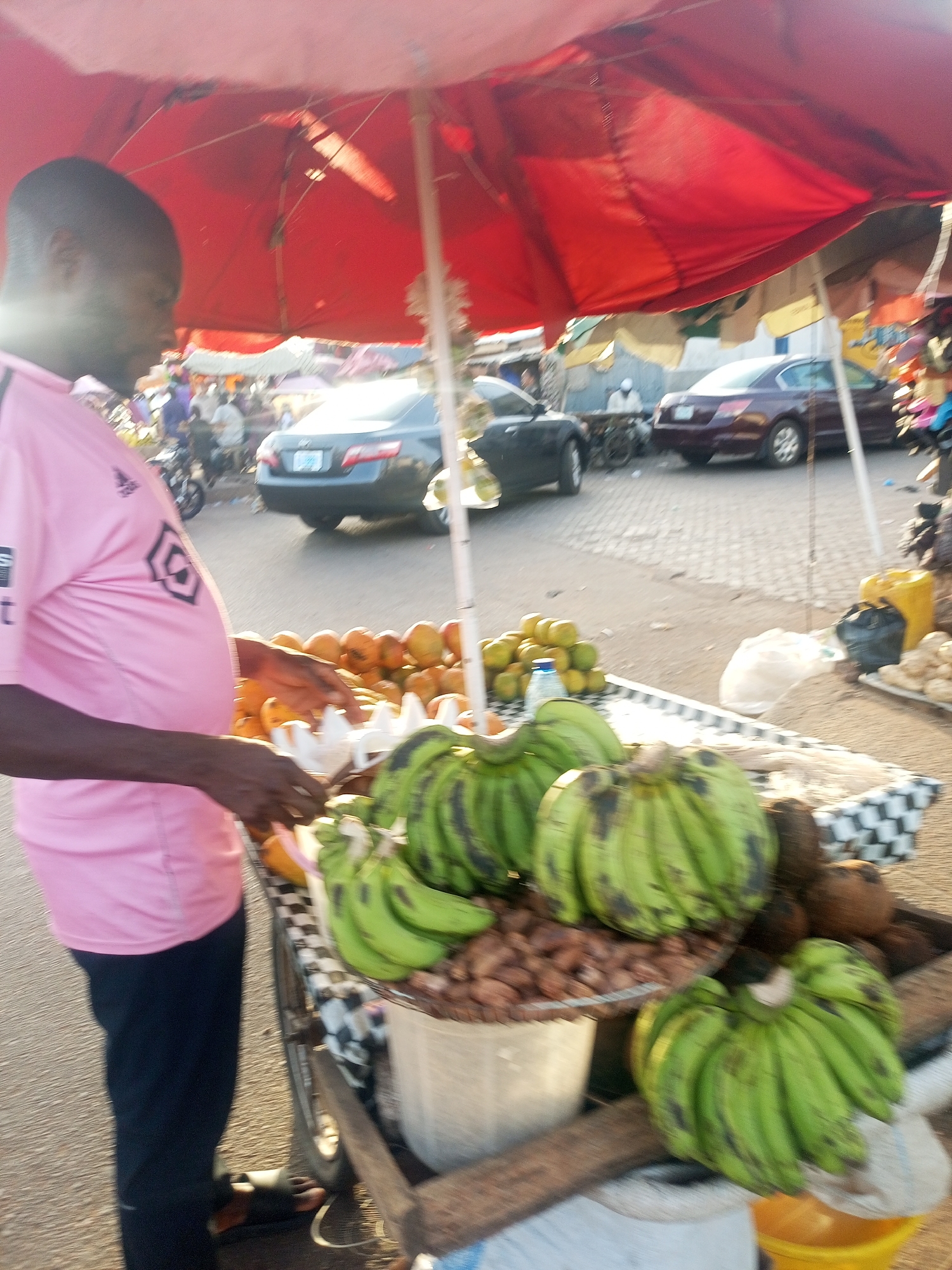 This image depicts an outdoor market scene in Tempure, Nigeria, likely during the late afternoon or early morning due to the warm, golden light hitting the ground and some objects.
In the foreground, a man in a pink t-shirt with a black hexagonal logo and dark pants stands beside a fruit stall. His profile is visible, but his face is somewhat obscured by shadow. He appears to be attending to the stall, possibly arranging produce or serving a customer, though no customer is visible.
The fruit stall is covered by a large red umbrella, providing shade from the sun. On the stall, there are several large bunches of green bananas or plantains, some showing dark spots. Piles of citrus fruits, possibly oranges or mangoes, are also displayed. Other items include a collection of brown nuts, likely kola nuts, in a container, and several brown coconuts. A white egg carton rests on the stall.
In the background, a street or market area is visible with cars, including a dark blue sedan and a grey sedan, and other faint figures of people and a bicycle or motorbike, indicating a lively, urban or semi-urban setting. The ground is paved, appearing to be a mix of cobblestones and asphalt. Near the stall, a yellow jerrycan and various plastic bags are visible. The overall scene portrays a typical bustling market environment.